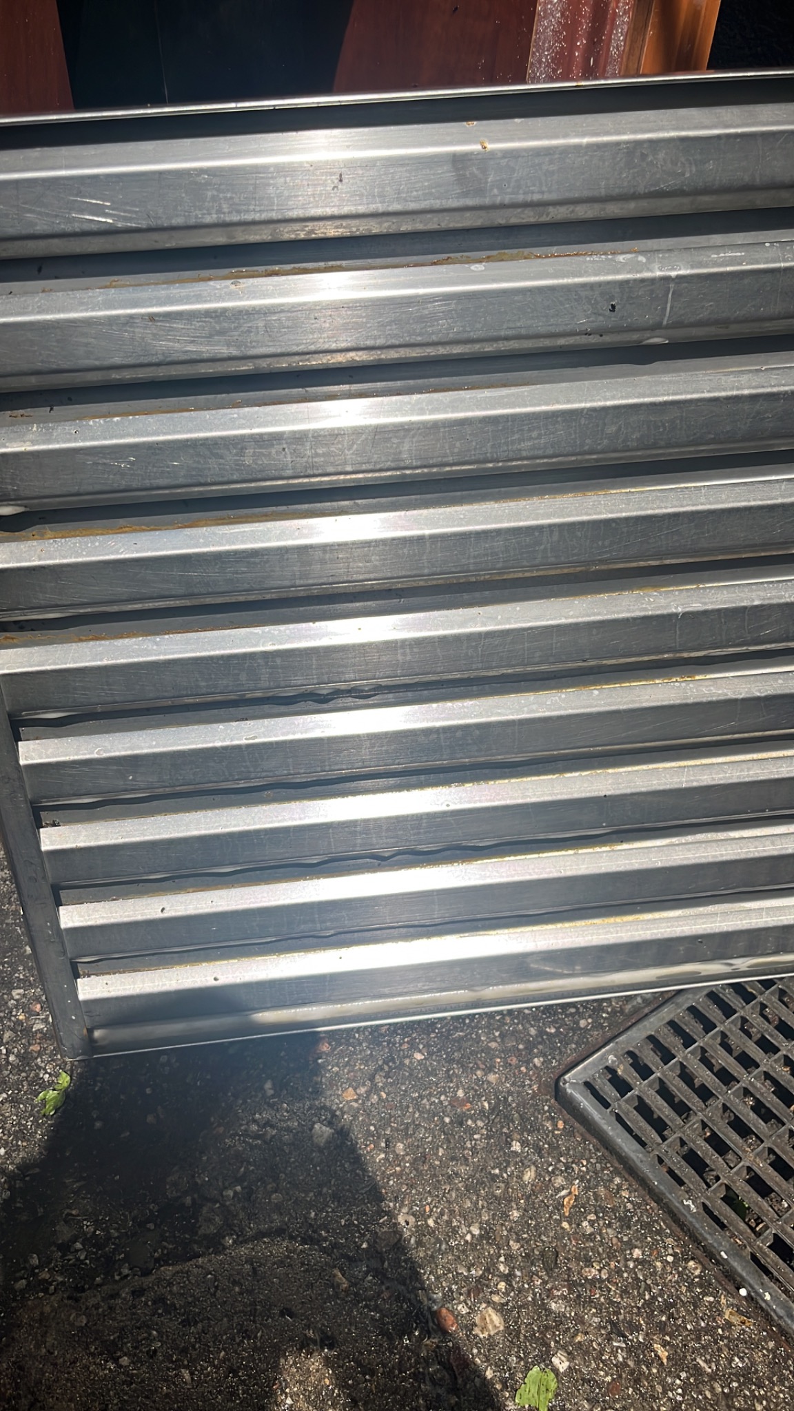 Stos metalowych żaluzji wentylacyjnych ułożonych na szarym chodniku obok kratki ściekowej.