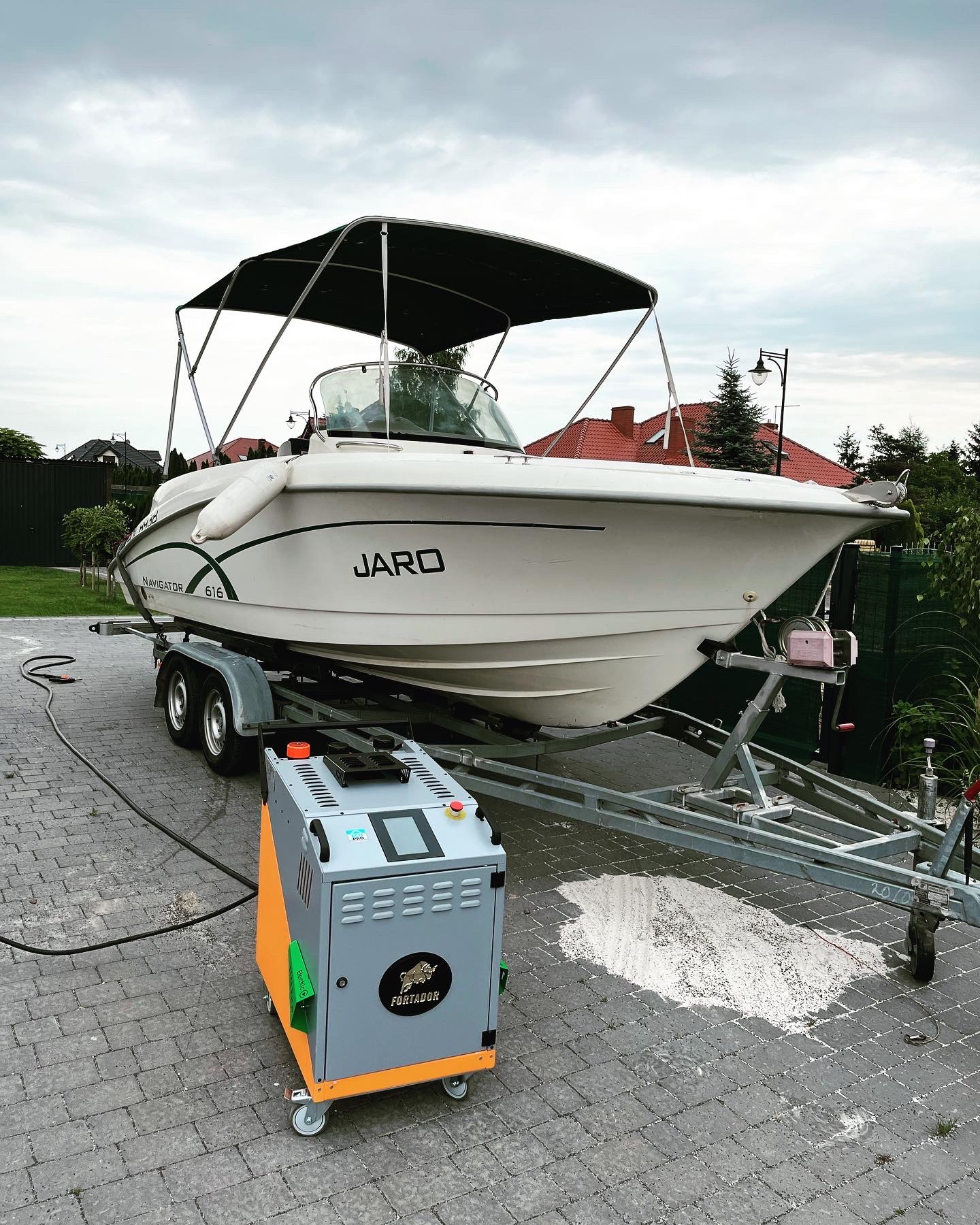 Biała łódź motorowa Navigator 616 z czarnym zadaszeniem, umieszczona na przyczepie, obok stoi szara myjka ciśnieniowa Fortador, widoczne ślady czyszczenia na brukowanej nawierzchni.