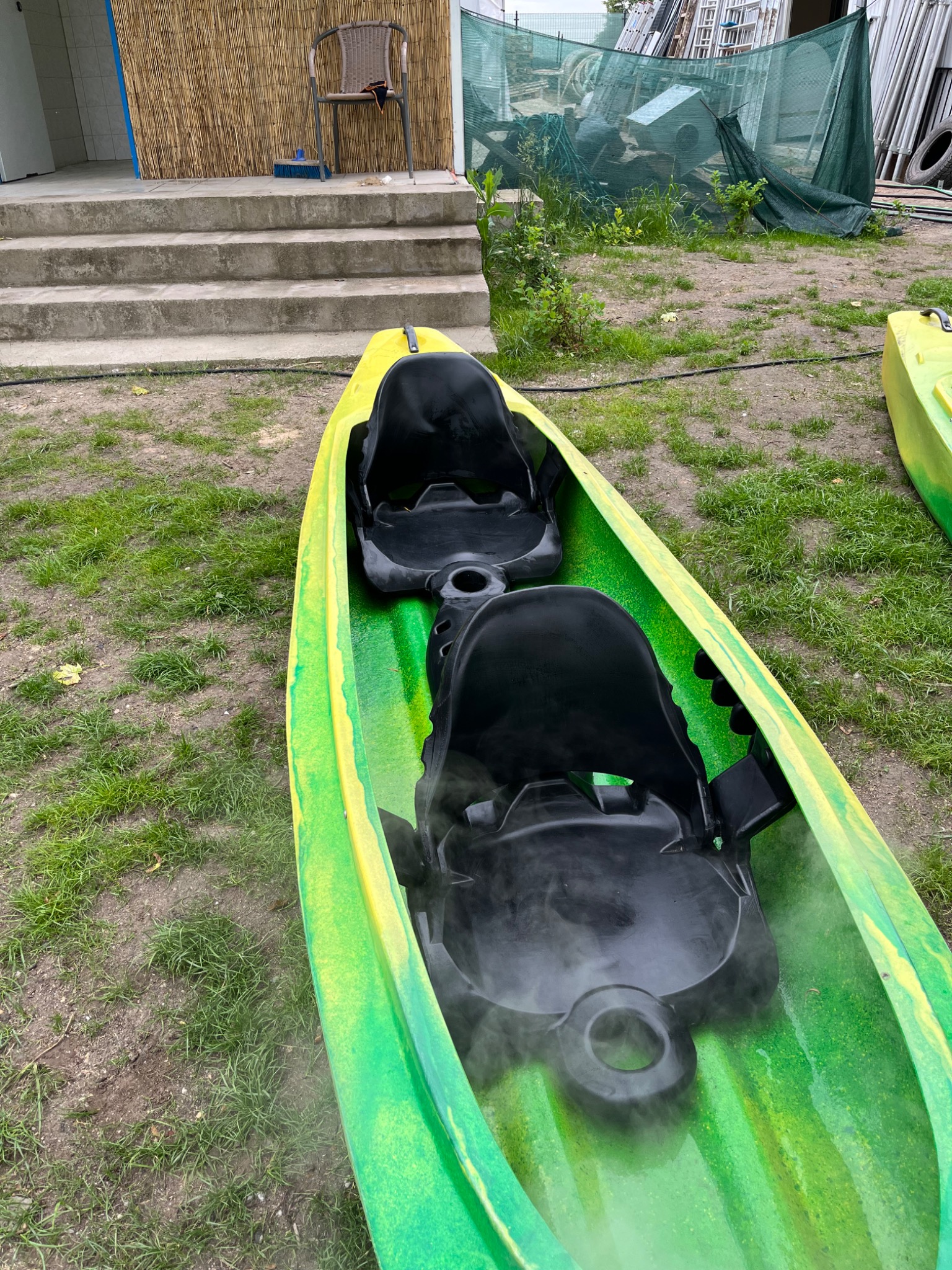 Zielono-żółty kajak dwuosobowy na trawie, z widocznymi kroplami wody na czarnych siedzeniach, sugerujący świeże umycie. W tle widoczne schody z bambusową zasłoną i zarys zagraconego podwórka.