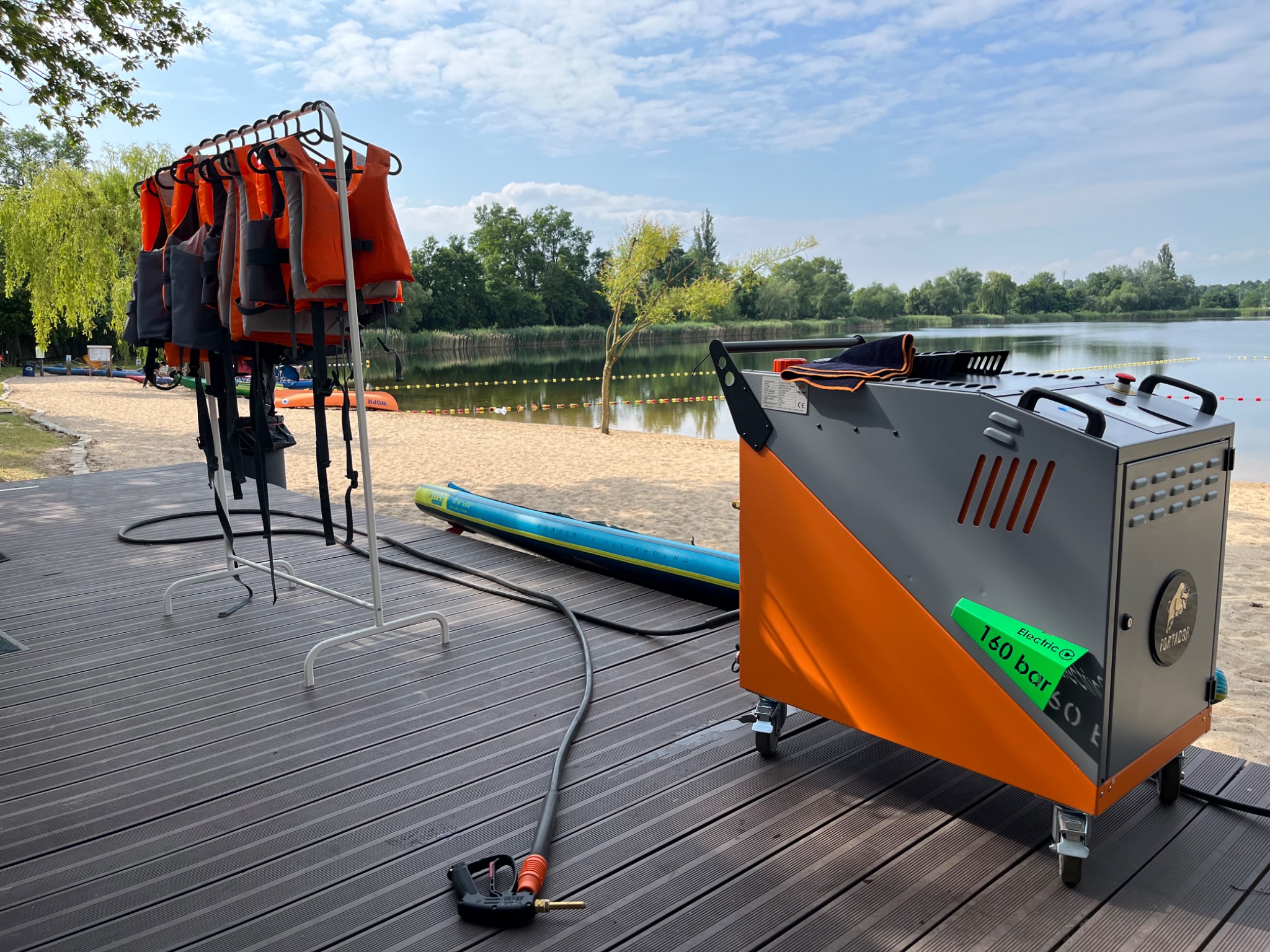 Mobilne urządzenie ciśnieniowe z dyszą, w tle wieszak z pomarańczowymi kamizelkami ratunkowymi, deska SUP na plaży nad jeziorem w słoneczny dzień.