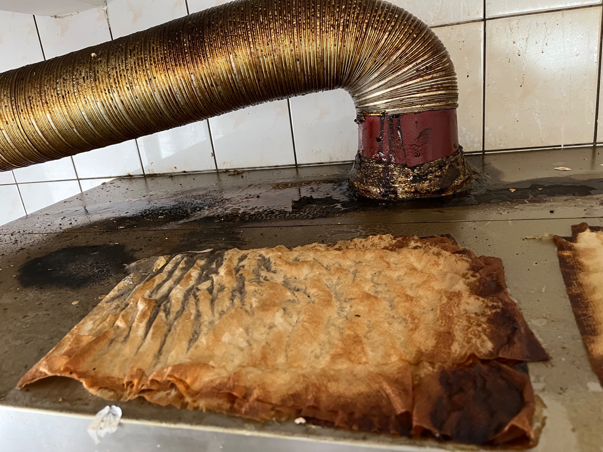 Zanieczyszczony, metalowy przewód wentylacyjny nad blatem roboczym z resztkami jedzenia i tłuszczu, widoczne białe płytki w tle.