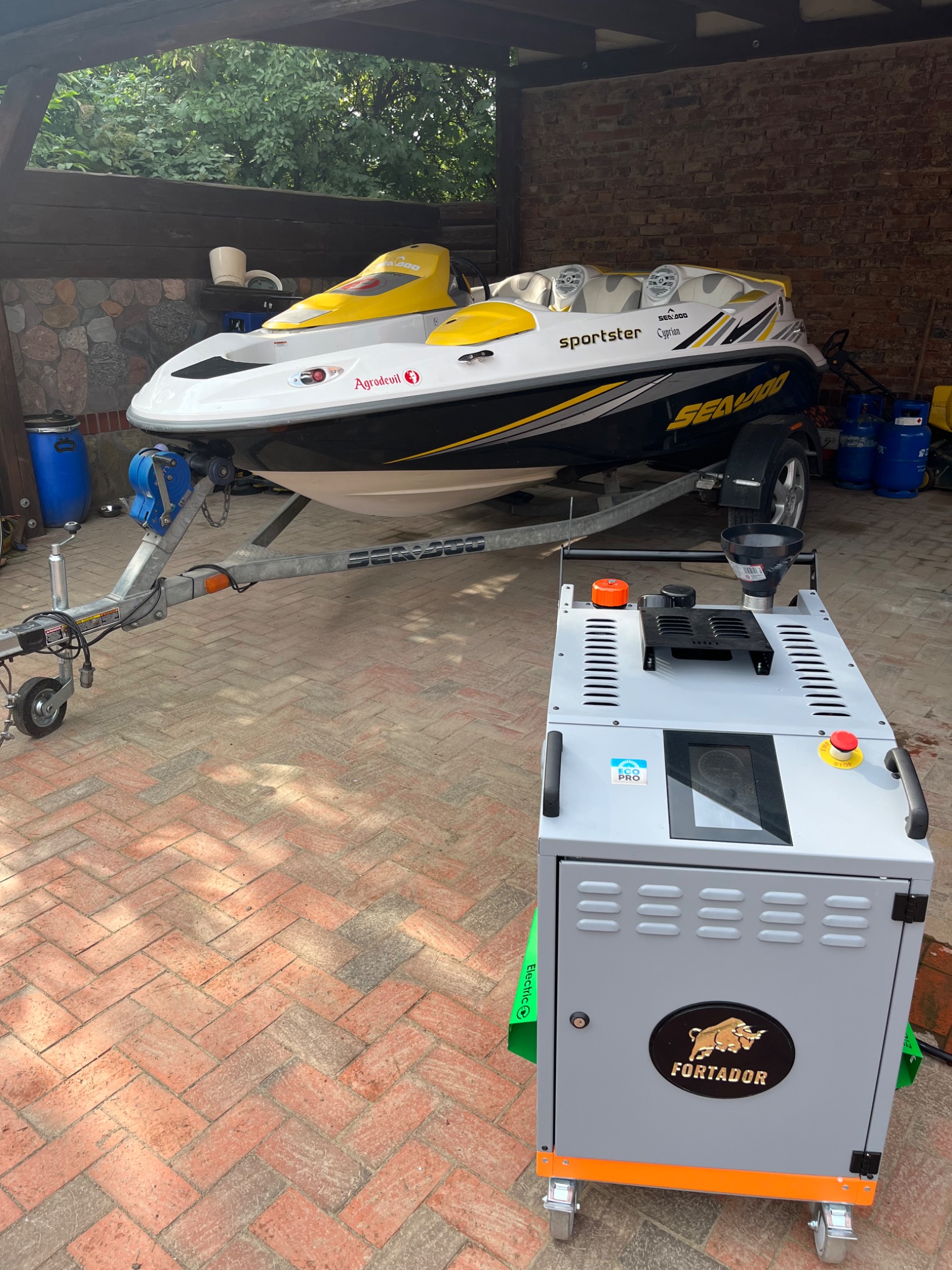 Żółto-biały skuter wodny Sea-Doo Sportster Cyprian na przyczepie, obok szarej maszyny czyszczącej Fortador z logo byka na ceglanym podłożu.