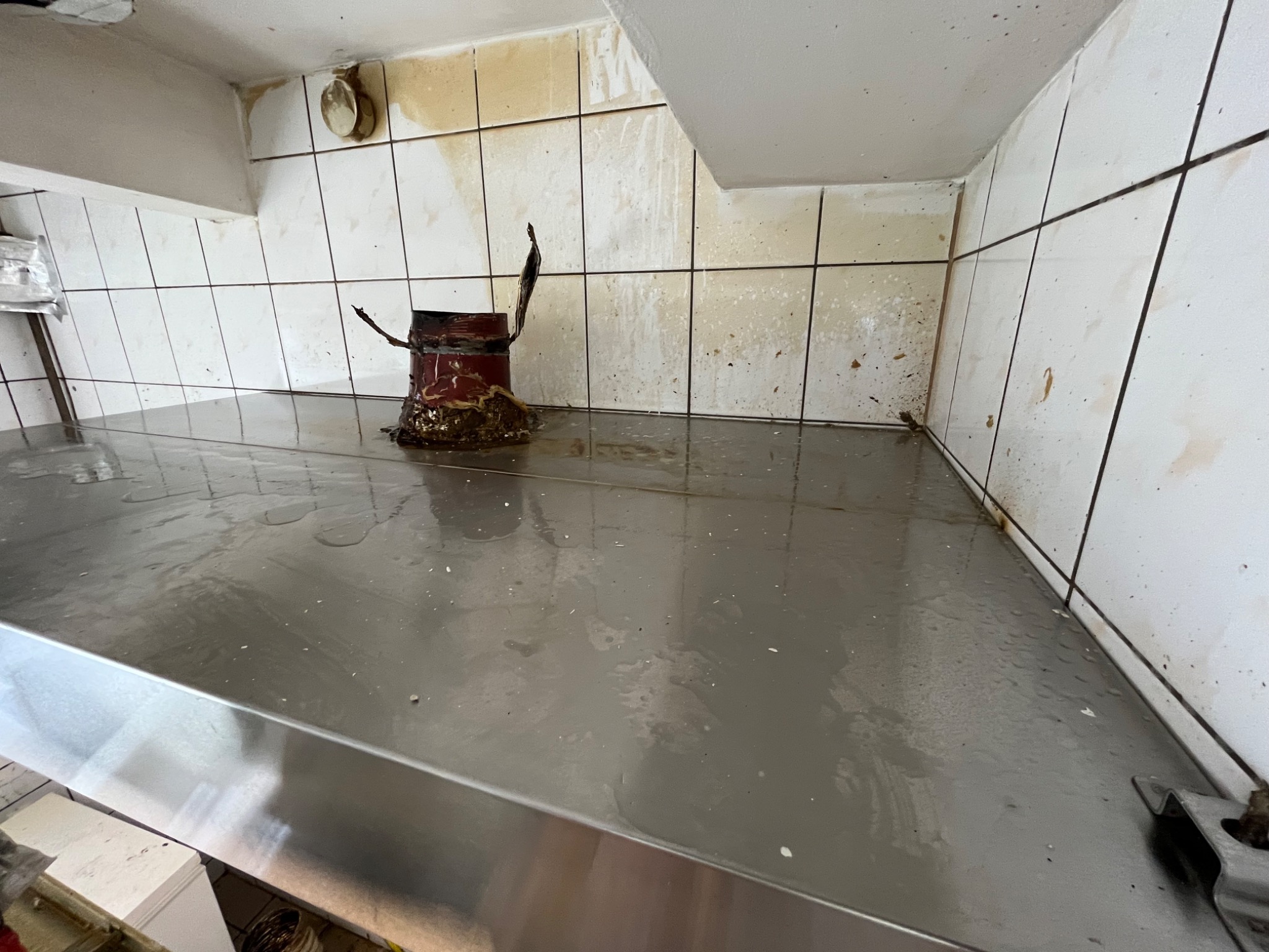 Zanieczyszczona powierzchnia robocza ze stali nierdzewnej w pomieszczeniu z brudnymi, białymi płytkami ceramicznymi na ścianach. Widoczny resztki po czyszczeniu lub zalaniu, oraz wiadro...