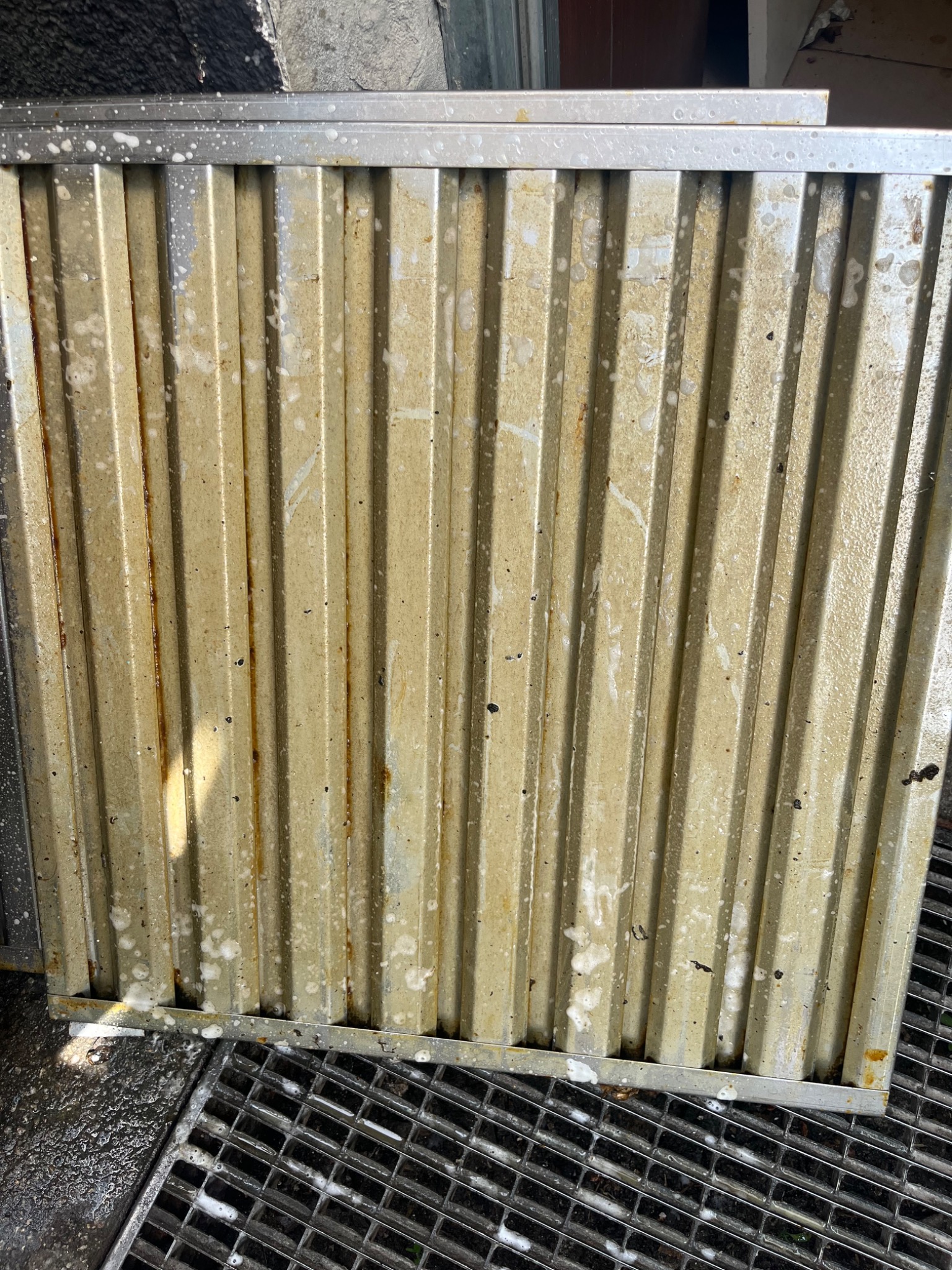 Brudny, żebrowany filtr przemysłowy z widocznymi śladami tłuszczu i resztkami detergentu, leżący na metalowej kracie ściekowej.