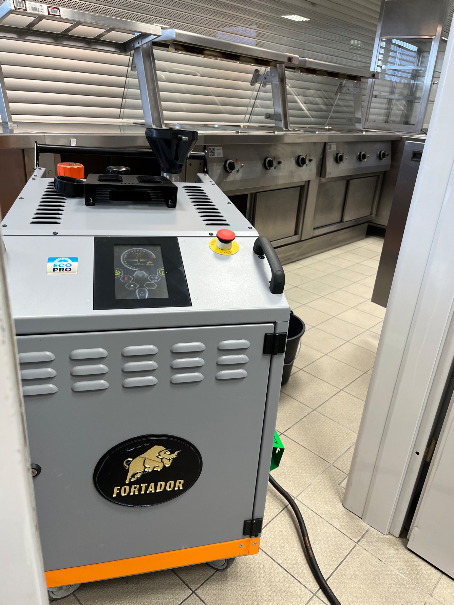 Szare urządzenie przemysłowe z czarnym lejem na górze, panelem sterowania z wyświetlaczem oraz logiem Fortador na dole, stojące na kafelkach w pomieszczeniu z metalowymi szafkami.