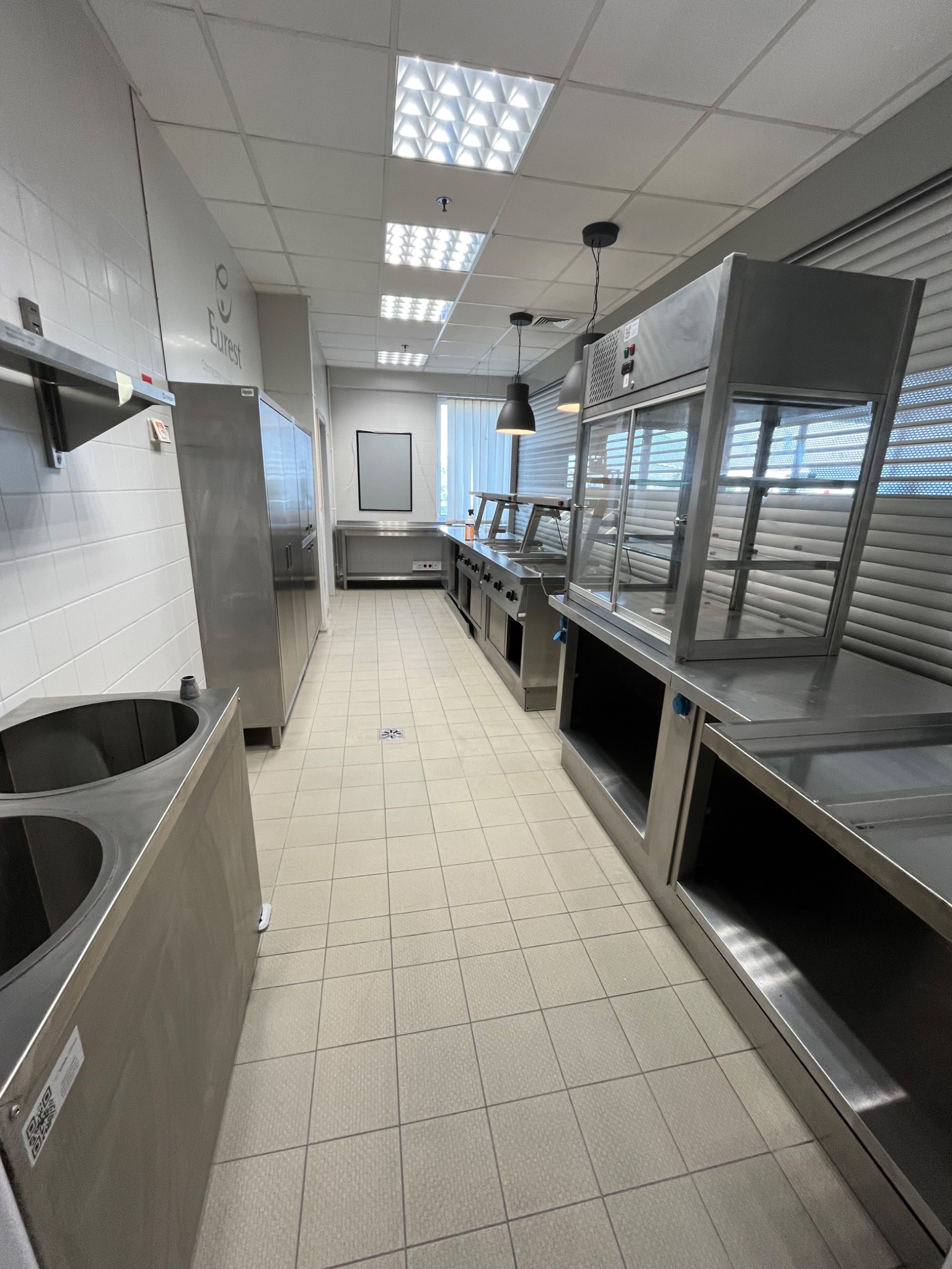 Wnętrze profesjonalnej kuchni z nierdzewnymi urządzeniami do serwowania posiłków, widok w głąb pomieszczenia z oświetleniem sufitowym i jasnymi kafelkami na podłodze.