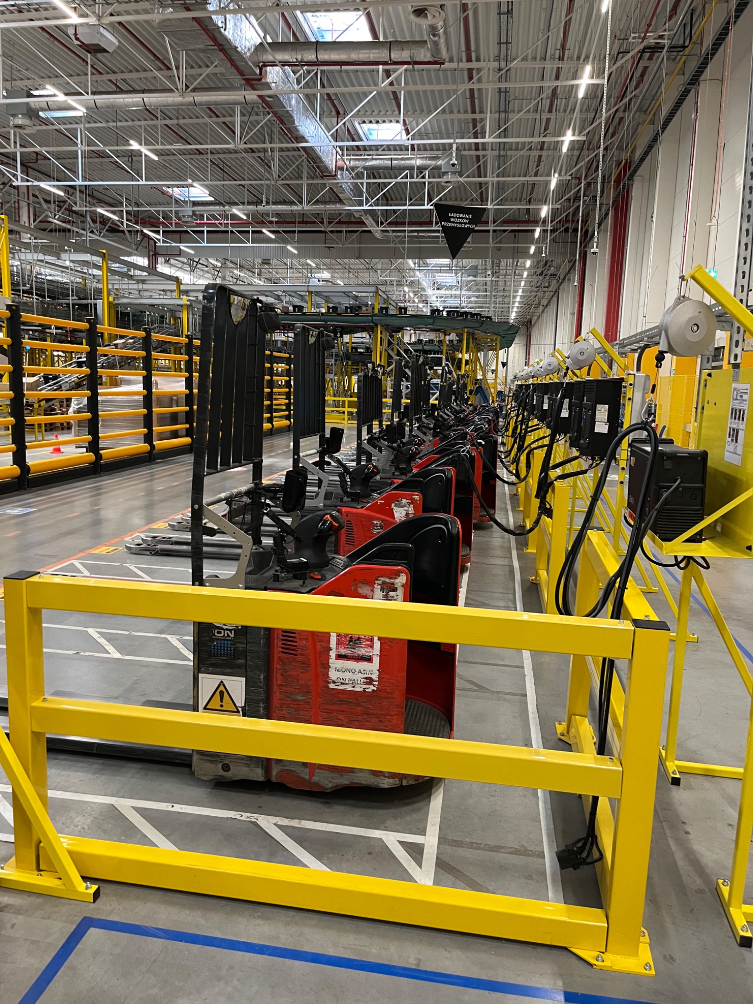 Rząd czerwonych wózków widłowych elektrycznych, zaparkowanych w hali produkcyjnej, zabezpieczonych żółtymi barierkami.