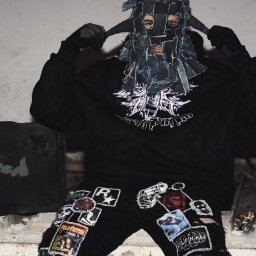 Michał Kapłanow mkvid media - Osoba w masce z denimu, czarnej bluzie i spodniach z naszywkami stoi w zrujnowanym wnętrzu obok czarnej walizki z napisem 'red'.