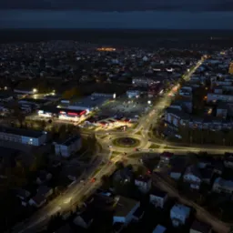 Nocny widok z drona na oświetlone miasto z okrągłym rondem, parkingiem i budynkami handlowymi.