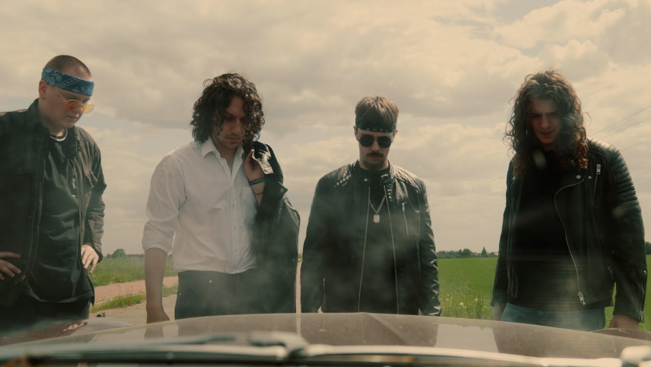 Czterech mężczyzn w stylizowanych strojach, stojących na drodze, uchwyconych z wnętrza samochodu, sugerujących scenę z filmu lub teledysku.