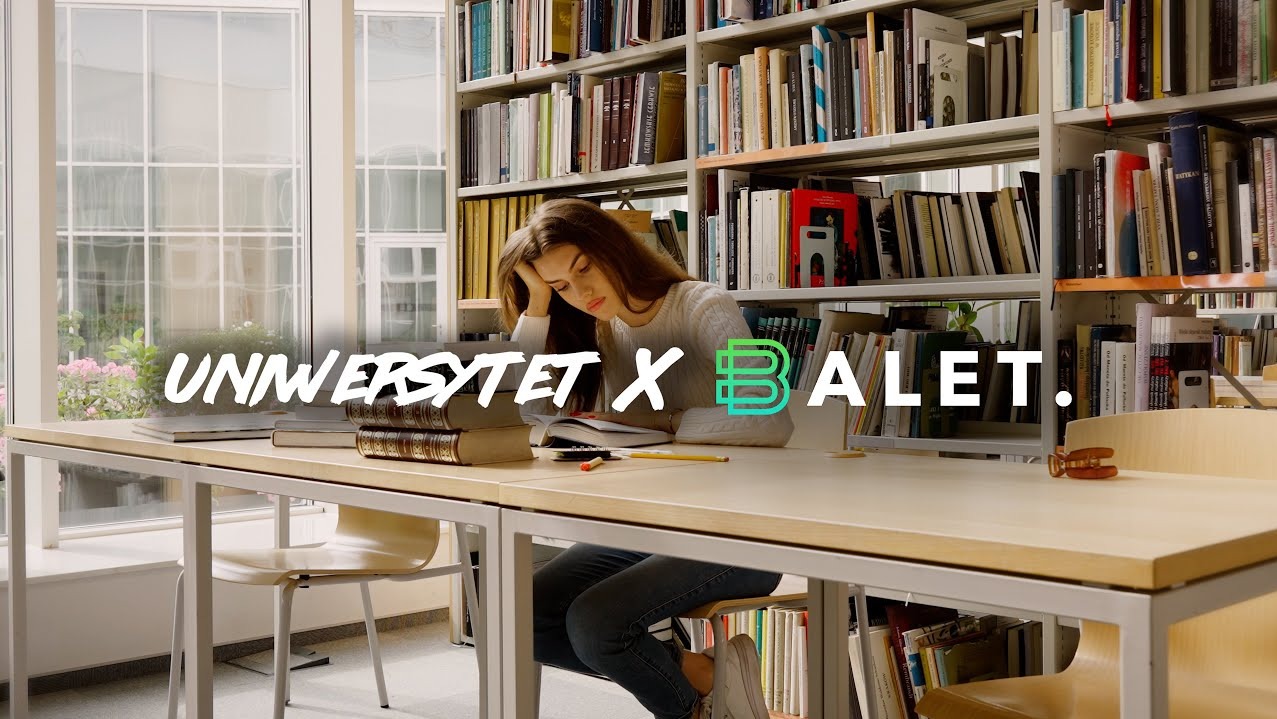 Kadr z filmu: studentka w bibliotece, czyta książkę przy biurku, w tle regały pełne książek i okno. Na pierwszym planie widoczne napisy i logo.