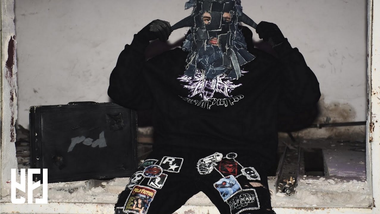 Osoba w masce z denimu, czarnej bluzie i spodniach z naszywkami stoi w zrujnowanym wnętrzu obok czarnej walizki z napisem 'red'.