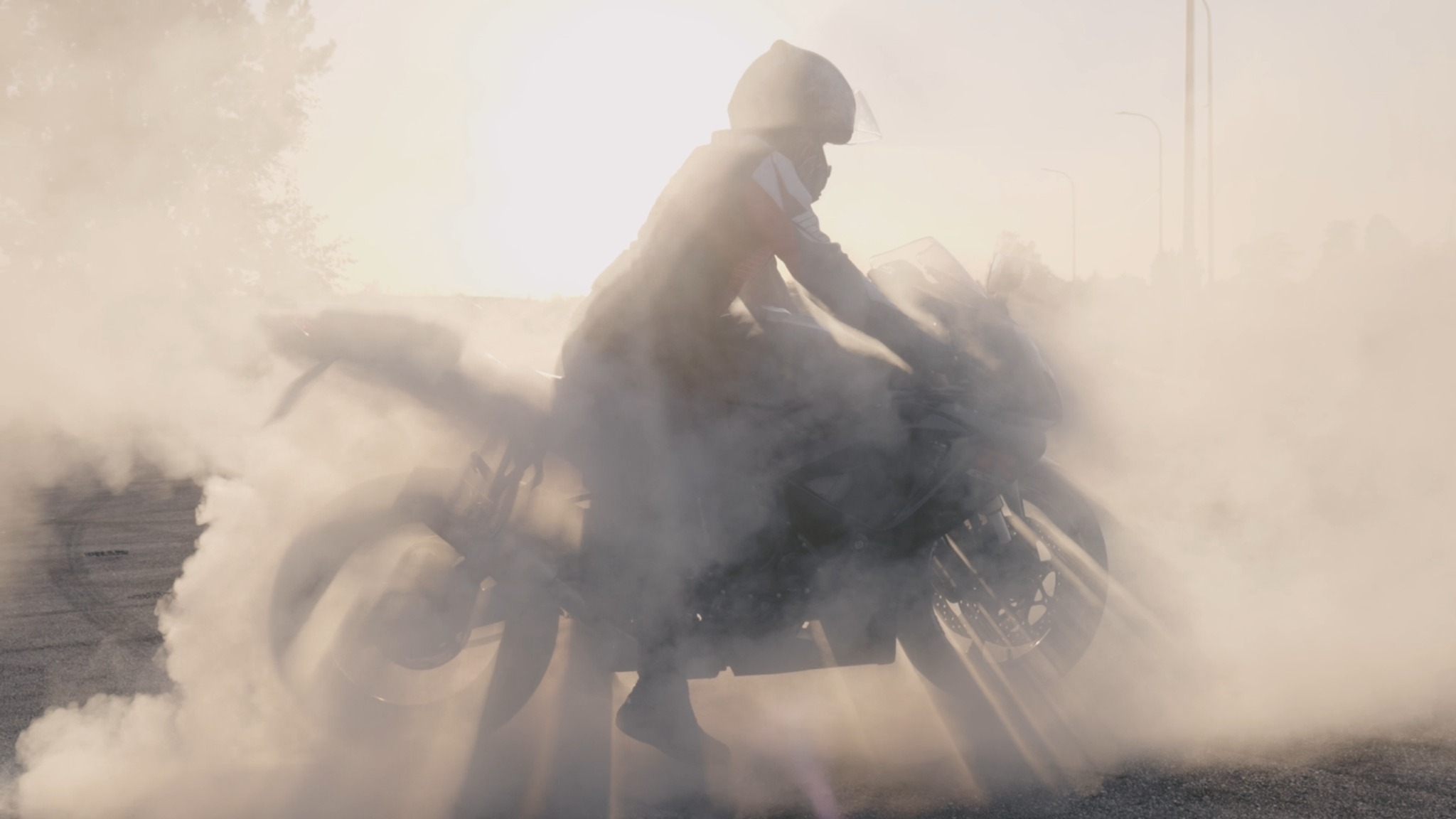 Motocyklista w kasku wykonuje burnout na motocyklu sportowym, gęsty dym zasłania otoczenie, widoczne oświetlenie uliczne w tle.