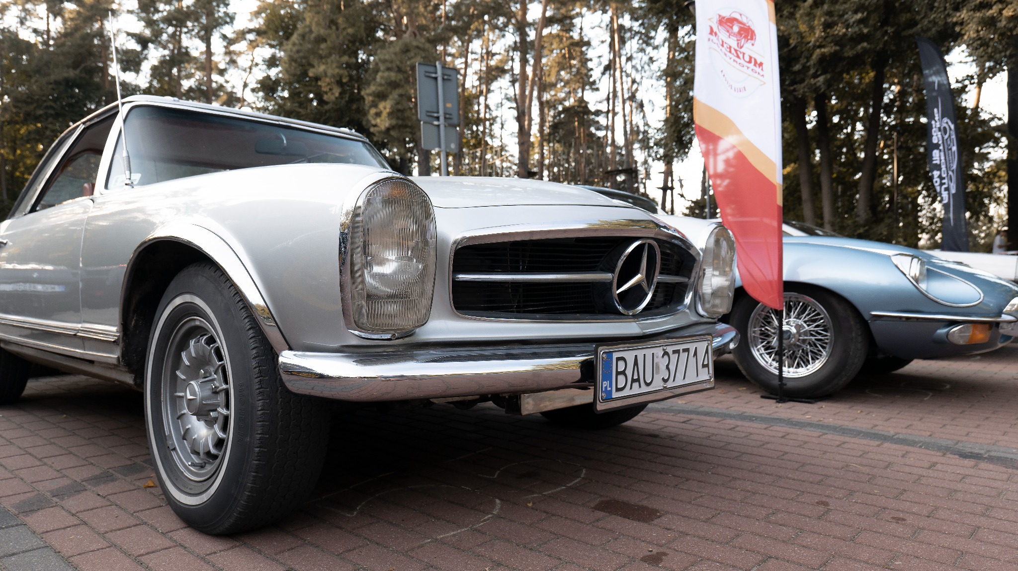 Zbliżenie na przód srebrnego Mercedesa SL z tablicą rejestracyjną z Białegostoku, zaparkowanego na czerwonej kostce brukowej, z widocznym fragmentem niebieskiego Jaguara E-Type w tle i flagami...
