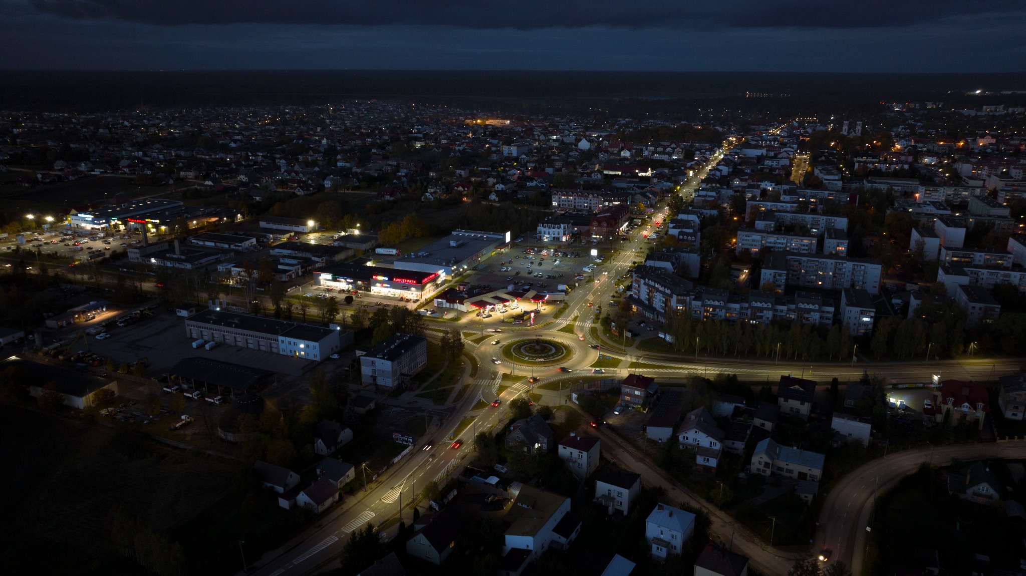 Nocny widok z drona na oświetlone miasto z okrągłym rondem, parkingiem i budynkami handlowymi.