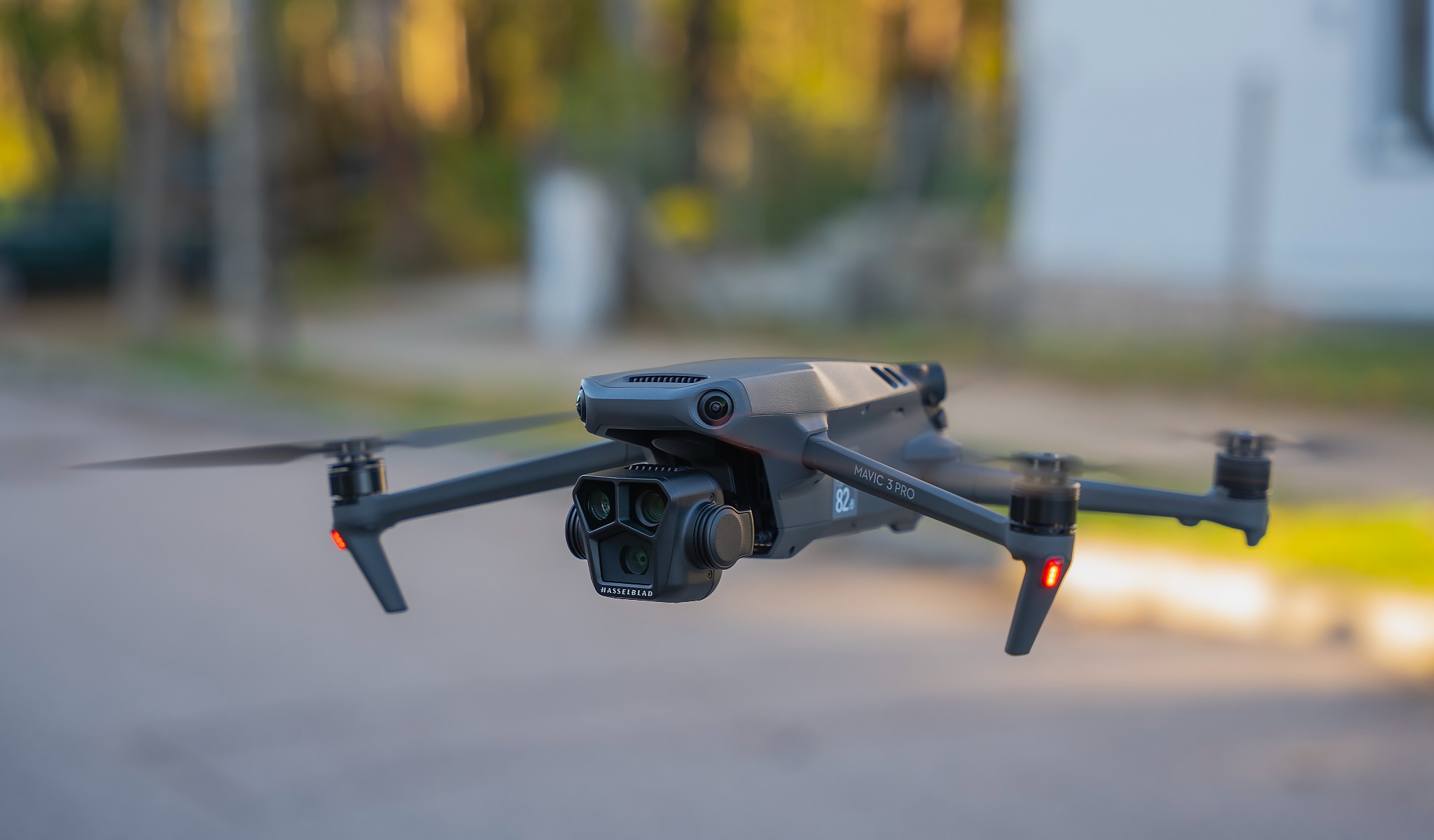 Szary dron DJI Mavic 3 Pro w locie na tle rozmytej zieleni i jasnej ściany budynku.