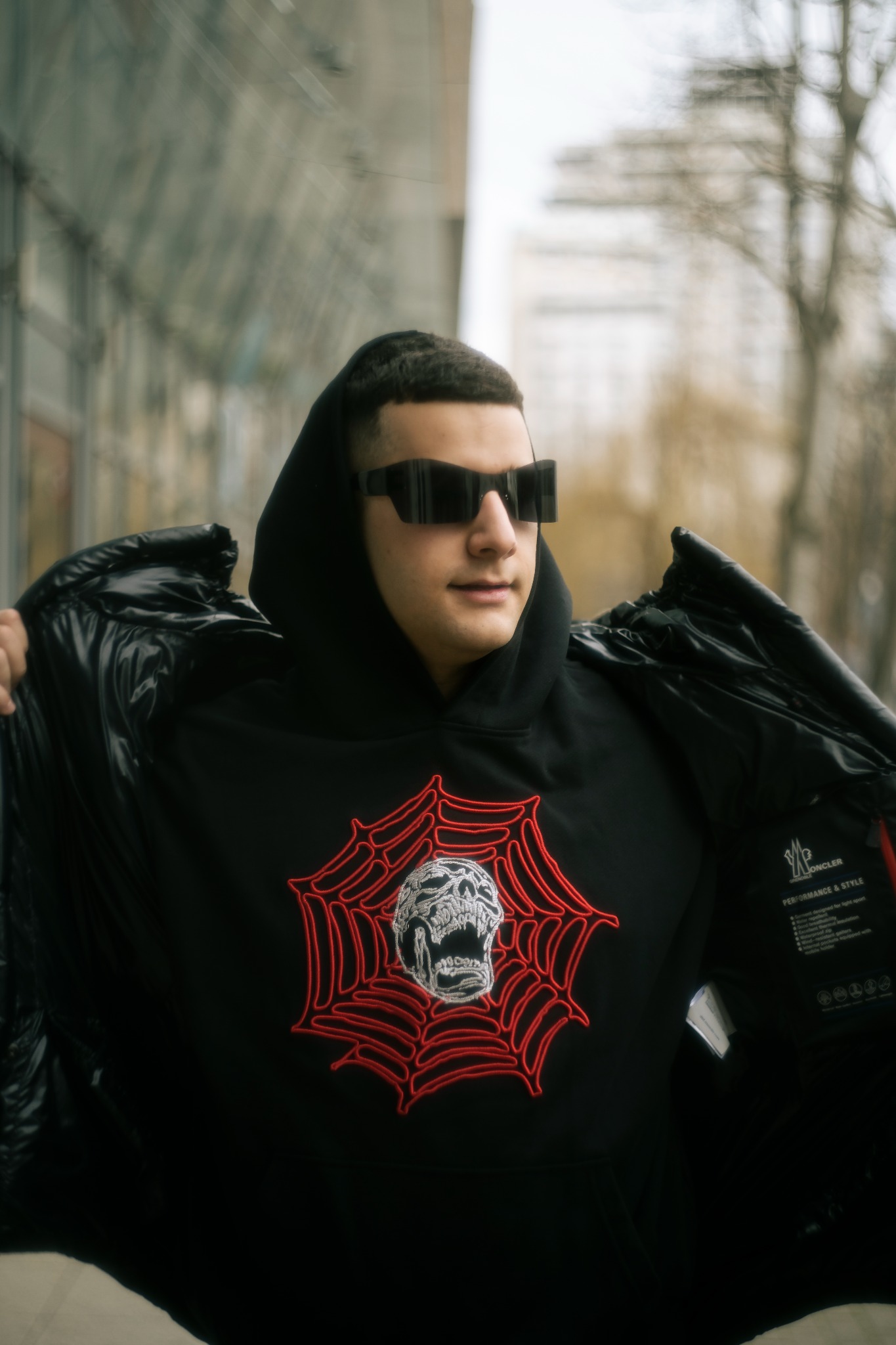 Mężczyzna w czarnej bluzie z kapturem z czerwonym motywem pajęczyny i czaszki, w czarnych okularach, trzymający rozpiętą czarną kurtkę puchową, na tle miejskiego krajobrazu.