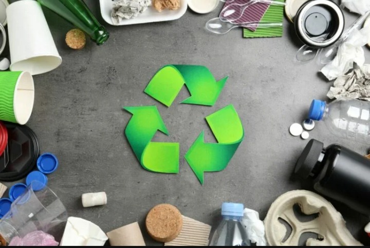Symbol recyklingu otoczony różnymi odpadami: plastikowymi butelkami, papierowymi kubkami, szklaną butelką i korkami, ułożone na szarym tle.