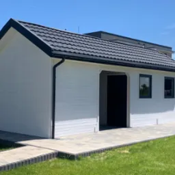 Nowoczesny, biały domek narzędziowy z czarnym dachem z blachodachówki, otoczony świeżo posianą trawą i wyłożony kostką brukową, w tle widoczne inne budynki.