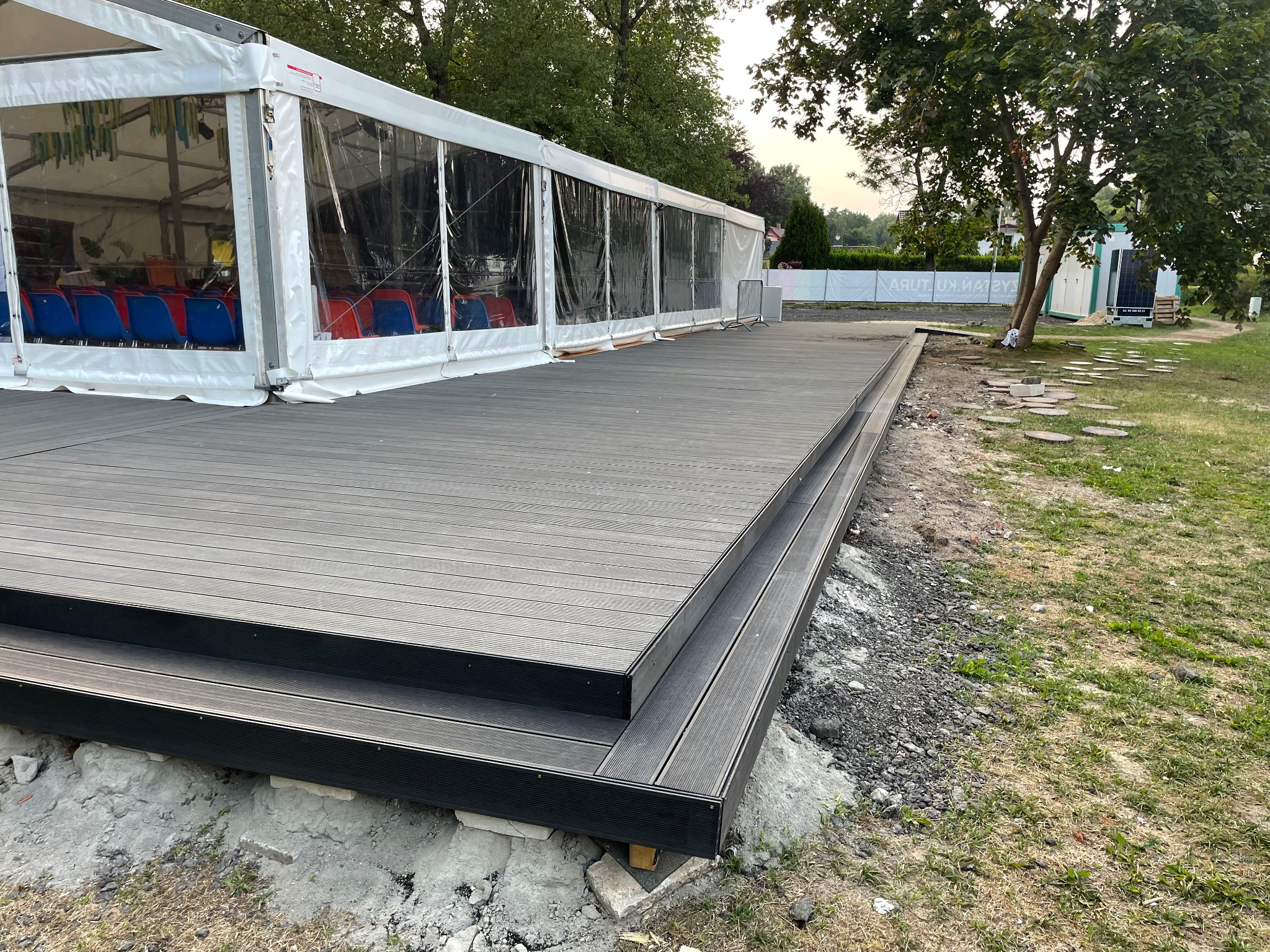 Nowoczesny taras z desek kompozytowych w kolorze grafitowym, z dwoma stopniami, przylegający do białego namiotu z przezroczystymi ścianami, w tle zieleń drzew i trawa, widoczne niedokończone prace...