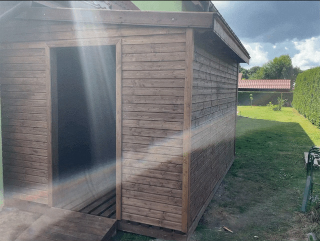 Drewniany domek ogrodowy z otwartymi drzwiami, widoczny fragment zadbanego trawnika i ogrodzenia.