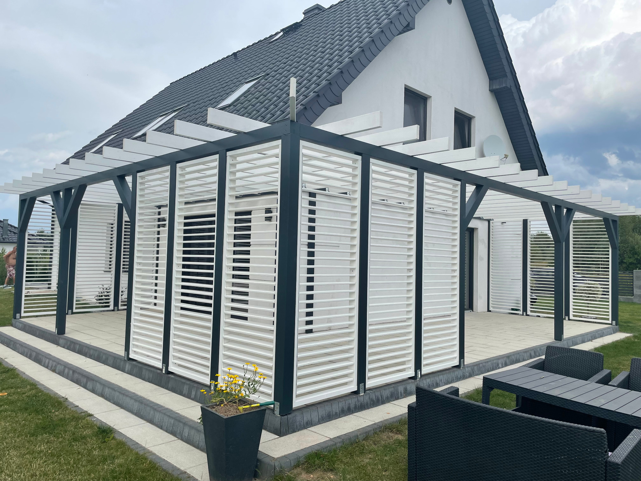 Nowoczesna pergola z białymi lamelami i ciemnoszarą konstrukcją, przylegająca do białego domu z ciemnym dachem, z widocznym zestawem mebli ogrodowych i donicą z żółtymi kwiatami.