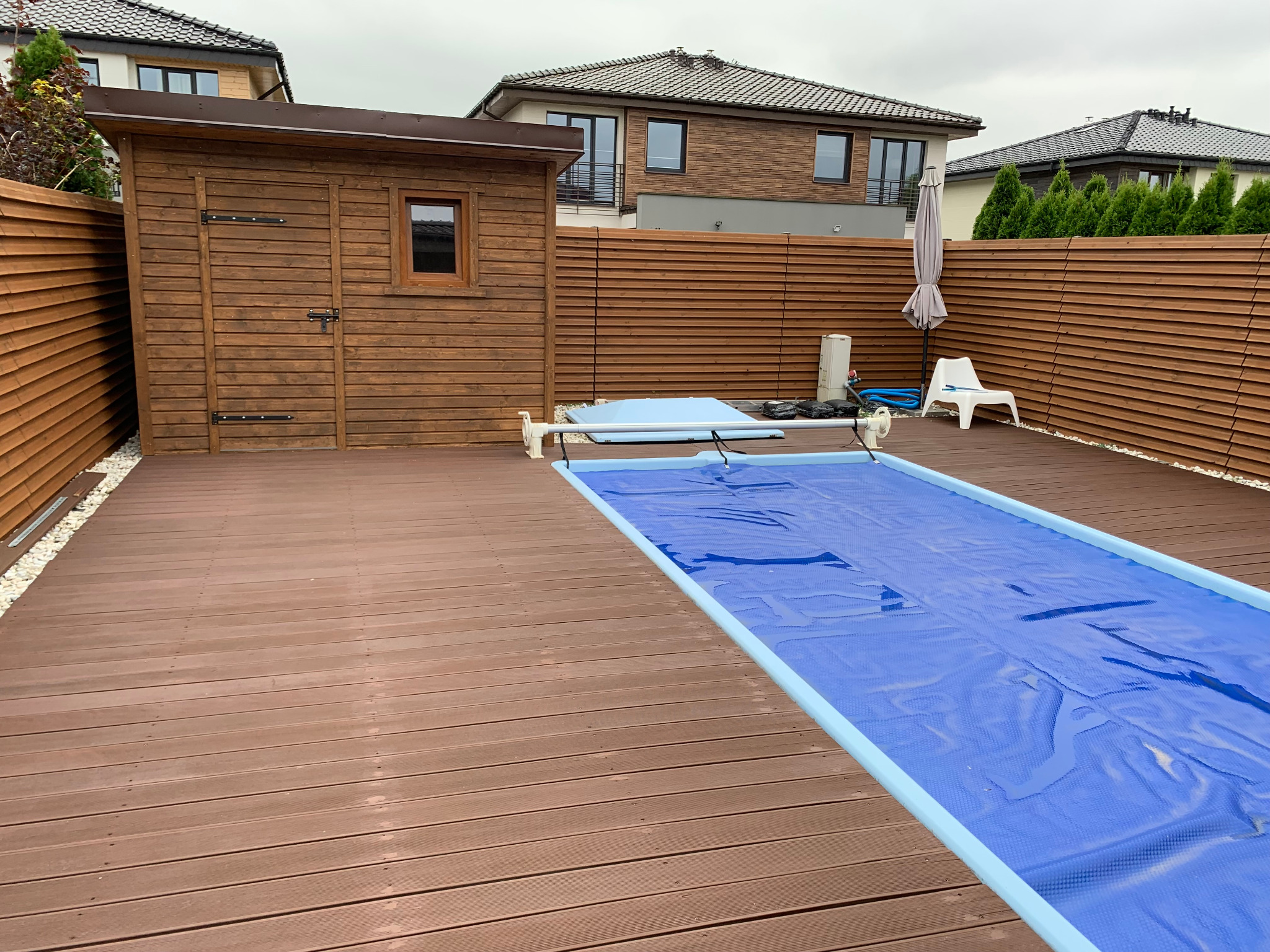 Zewnętrzny basen przykryty niebieską folią na drewnianym tarasie, obok brązowa drewniana szopa i wysoki płot z poziomych desek, w tle domy.