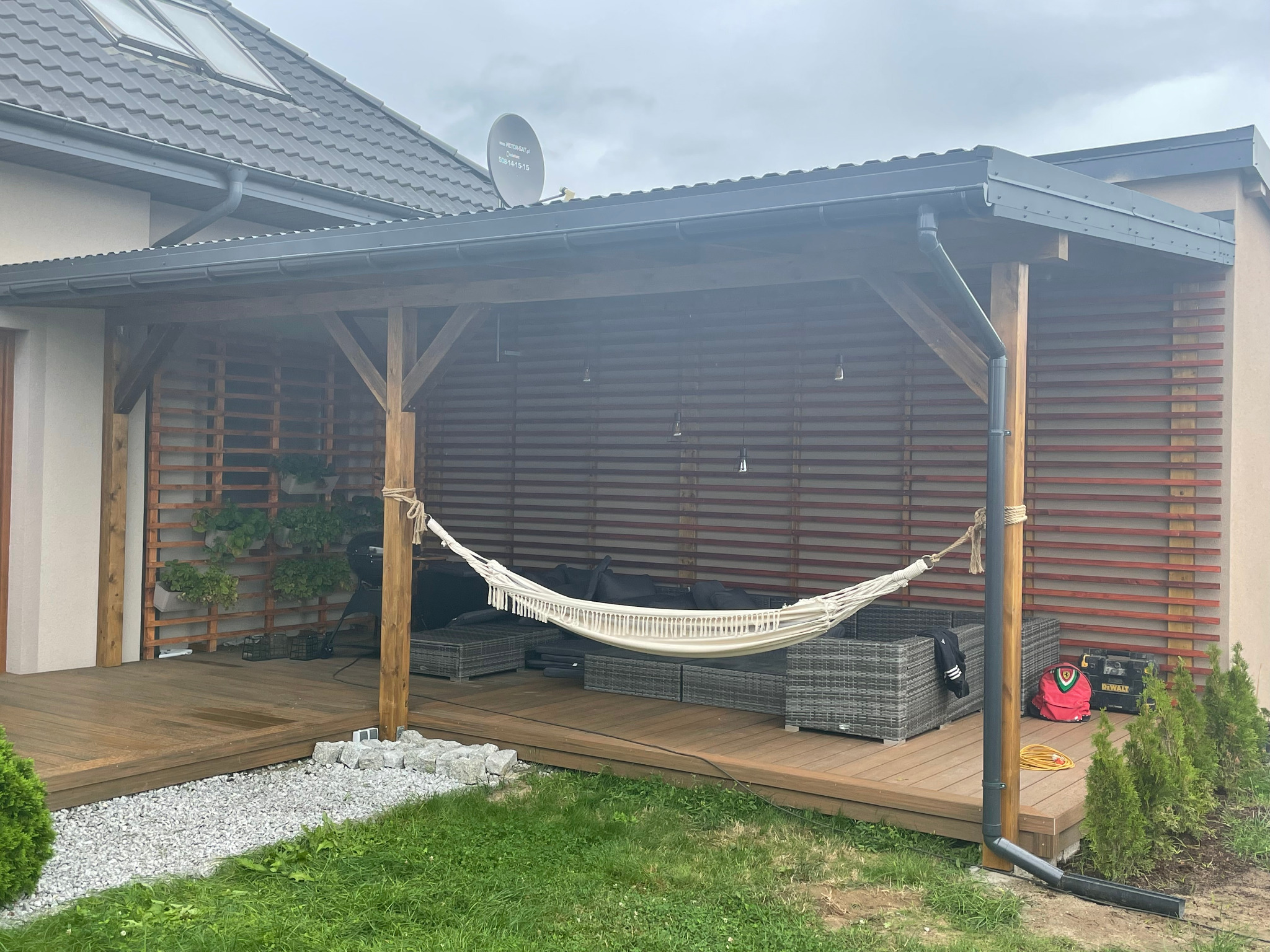 Zadaszony taras z drewnianą konstrukcją, hamakiem, meblami ogrodowymi z rattanu, pionową ścianą z roślinami w doniczkach i fragmentem trawnika z młodymi tujami