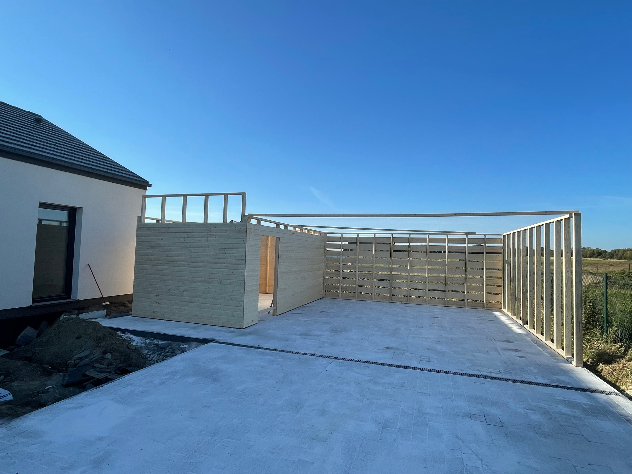 Budowa drewnianej konstrukcji na betonowej nawierzchni, z widoczną częścią domu i błękitnym niebem w tle, w trakcie wznoszenia ścian i zadaszenia.