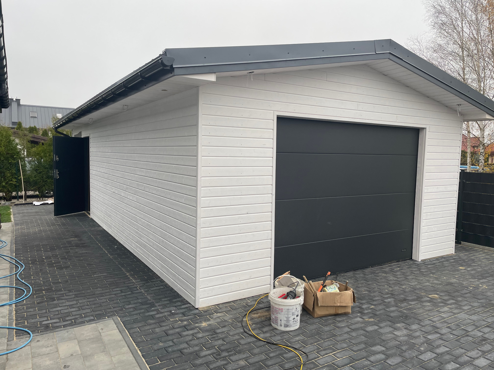 Nowo wybudowany, biały, drewniany garaż z szarą bramą segmentową, otwartymi drzwiami bocznymi, narzędziami malarskimi i wiaderkiem farby na szarej kostce brukowej.