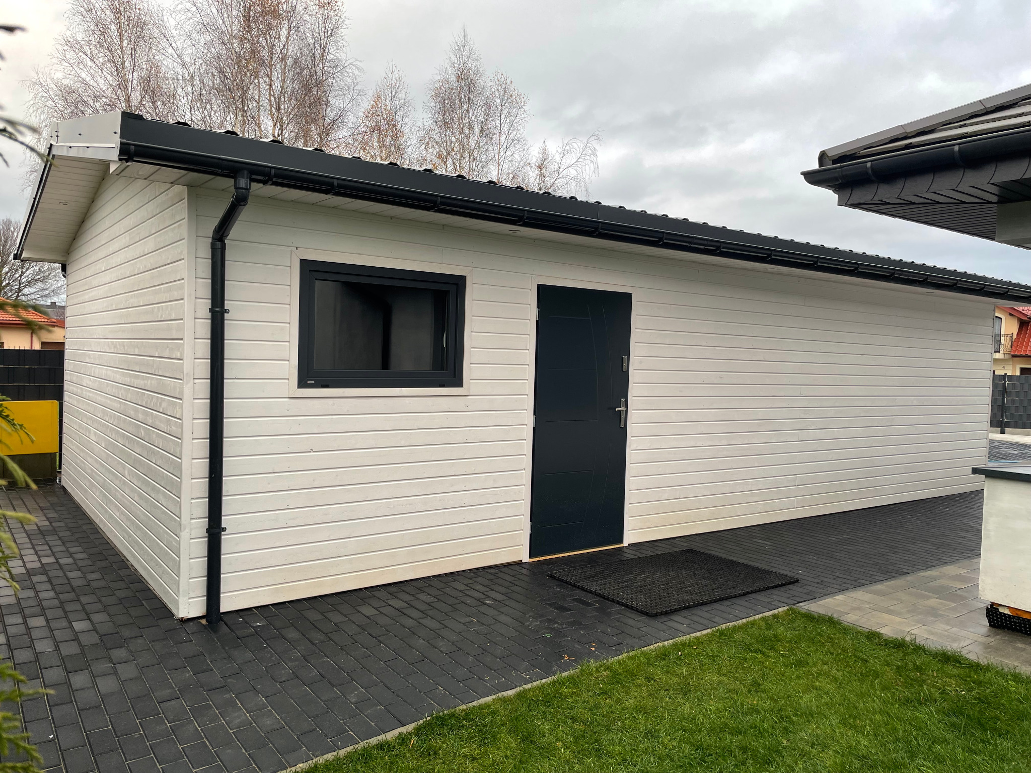 Nowoczesny, biały budynek gospodarczy z czarnymi detalami: okno, drzwi, rynny. Budynek otoczony kostką brukową i trawnikiem. Widoczne drzewa w tle.