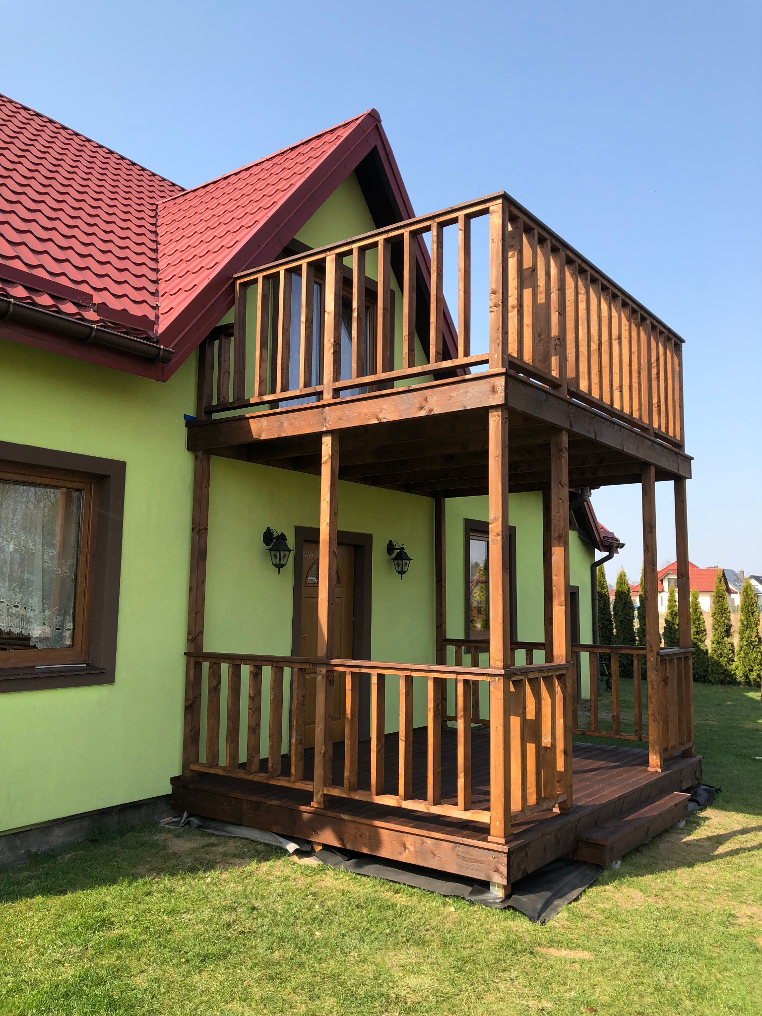 Dwukondygnacyjny, drewniany taras z balustradą przy domu z zieloną elewacją i czerwonym dachem, widok z trawnika w słoneczny dzień.