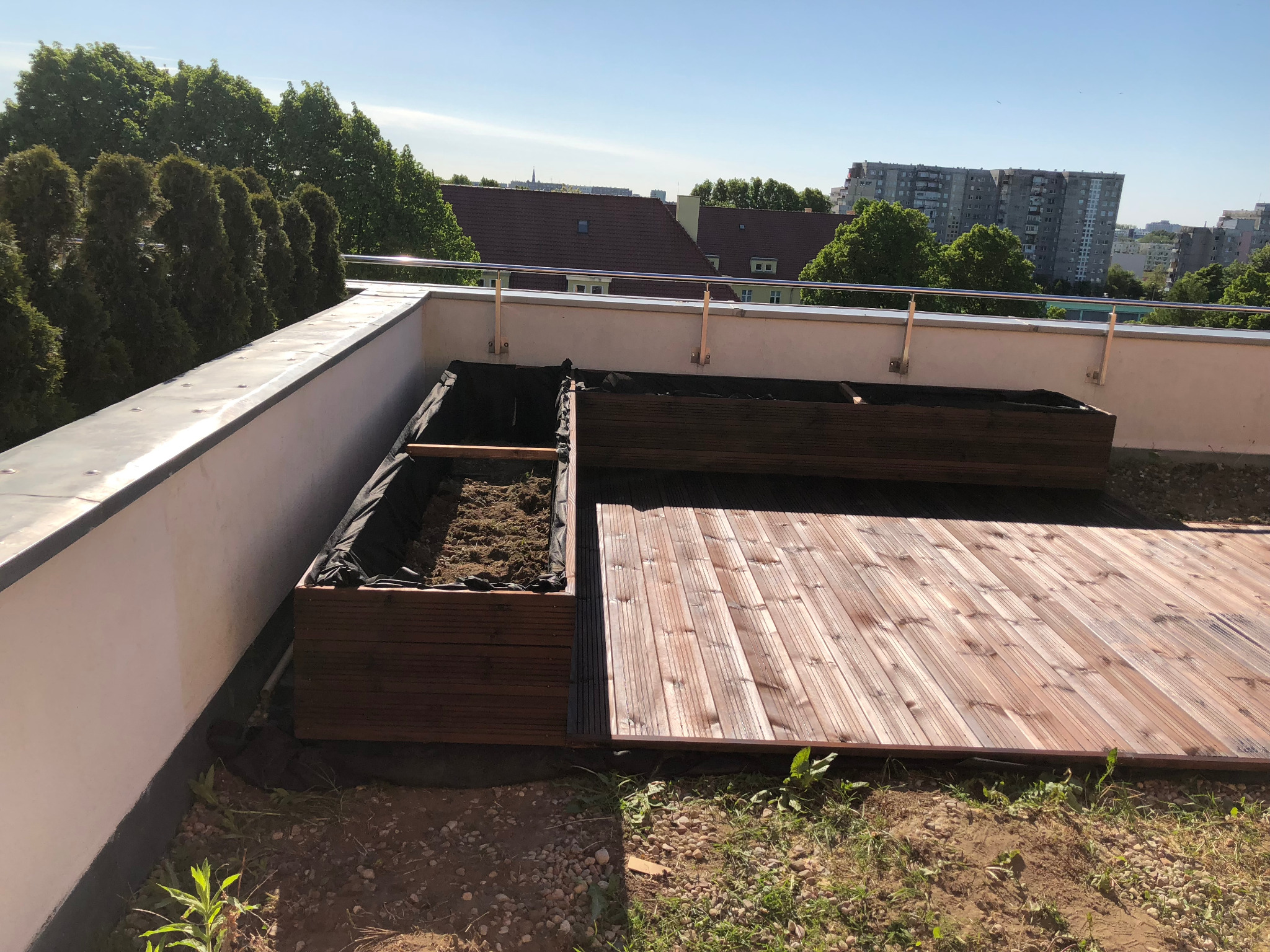 Drewniane skrzynie wypełnione ziemią na tarasie z desek, widok na panoramę miasta w tle.