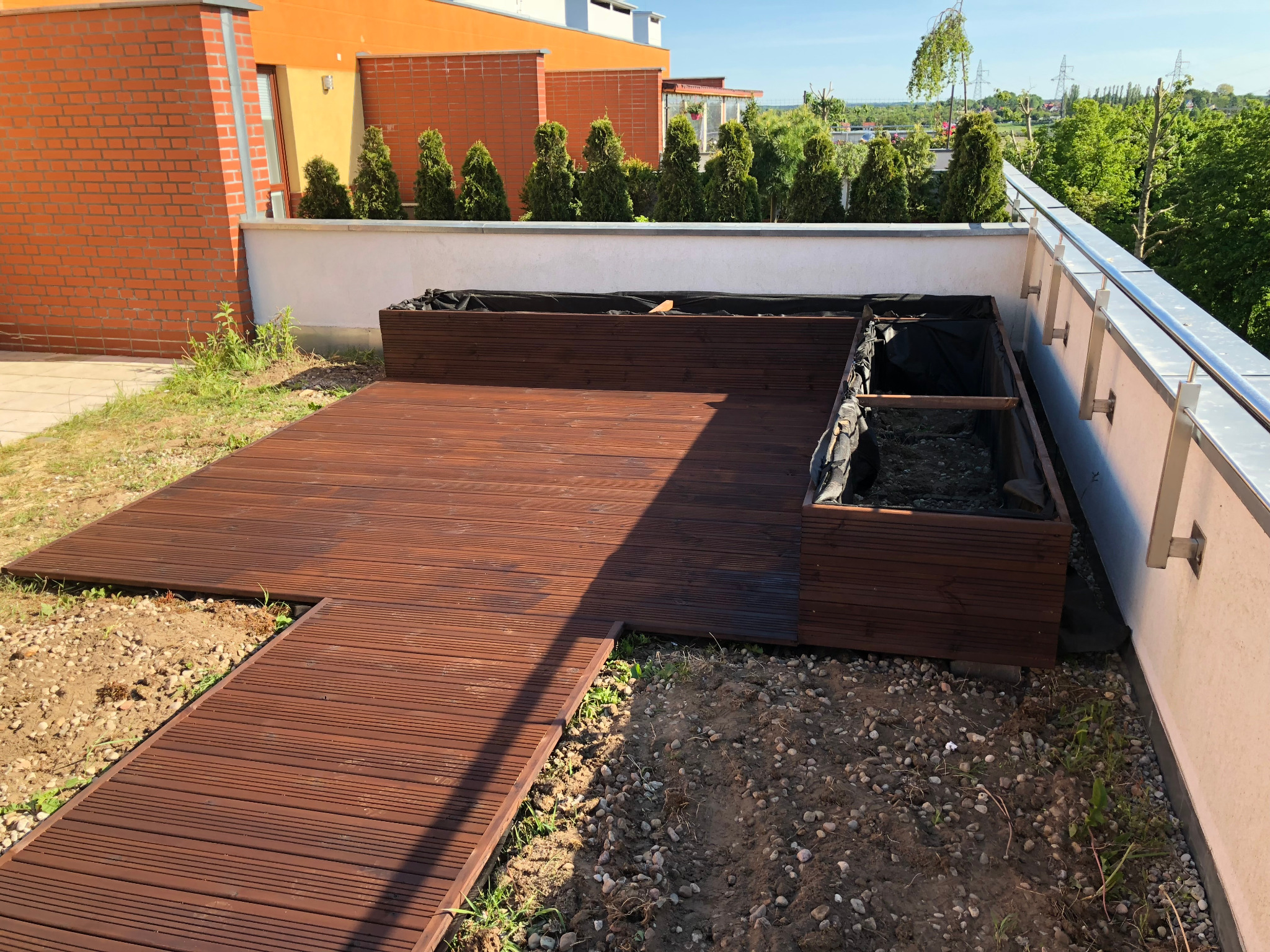 Drewniany taras na dachu budynku z przygotowanymi skrzyniami na rośliny, widok na zieleń i budynki w tle.