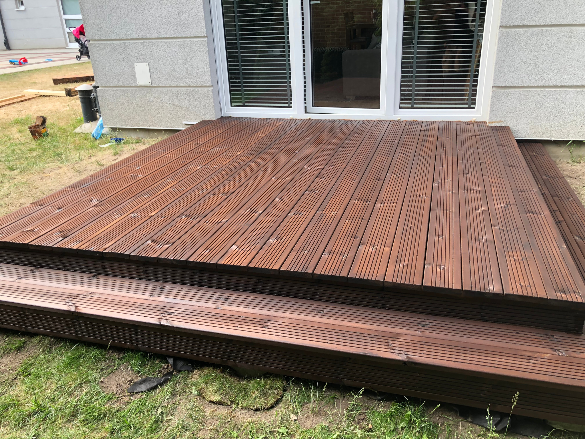 Nowo wybudowany, drewniany taras z ryflowanych desek w odcieniach brązu, przylegający do białej ściany domu z dużym oknem tarasowym. Widoczne schodki tarasowe i trawa wokół. W tle wózek dziecięcy...