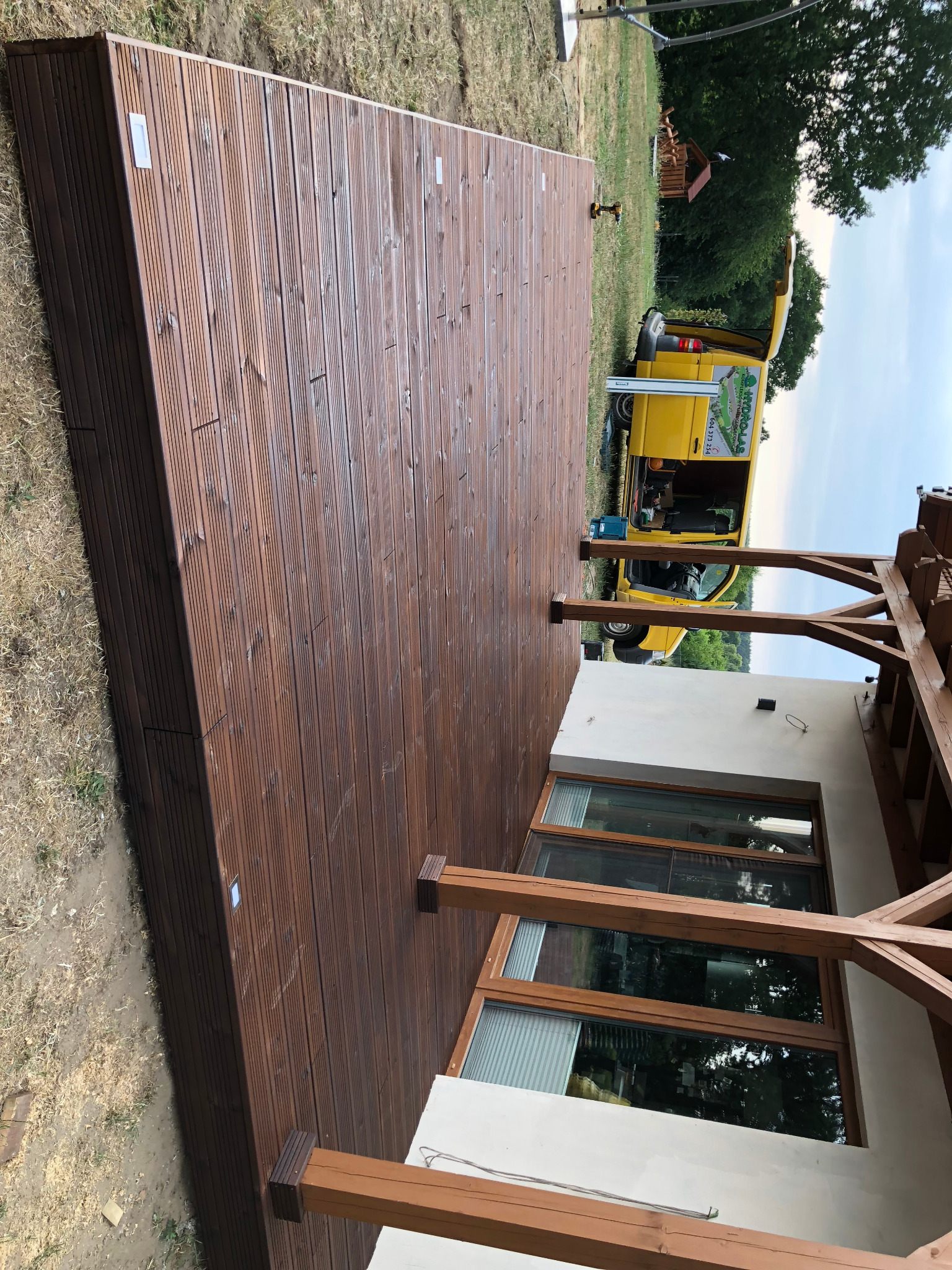 Nowoczesny taras z brązowych desek kompozytowych, przylegający do białego budynku z dużymi oknami, w tle żółty samochód dostawczy z otwartymi drzwiami bagażnika.