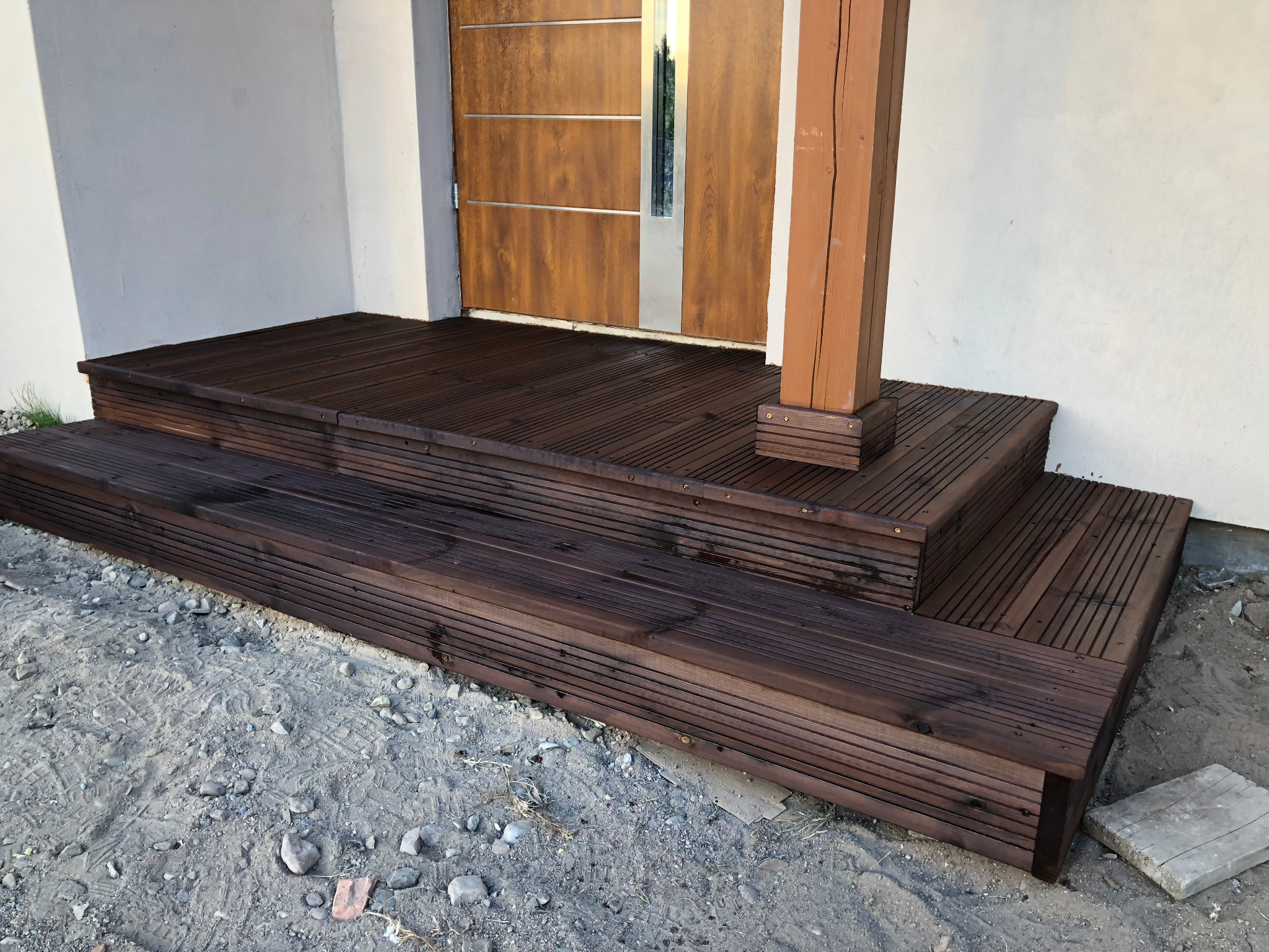 Nowoczesne drewniane schody wejściowe z podestem, prowadzące do drzwi z elementami stali nierdzewnej, osadzone na gruncie z piaskiem i kamieniami, z widoczną drewnianą kolumną wspierającą zadaszenie.