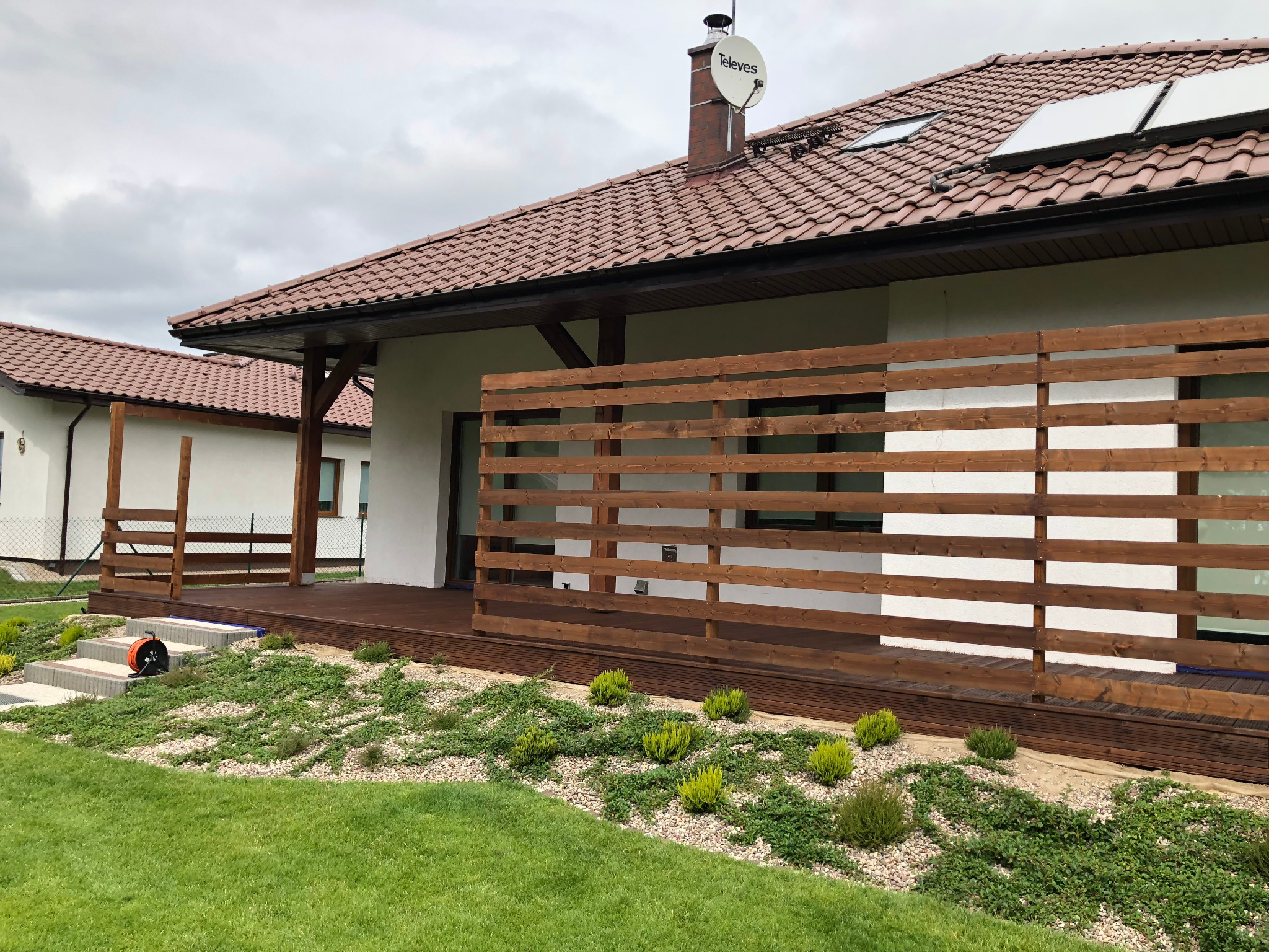 Nowoczesny ogród przydomowy z drewnianym tarasem i ażurowym ogrodzeniem, ozdobne kamienie, niska roślinność okrywowa i trawnik, widok na dom z brązową dachówką i panele słoneczne.