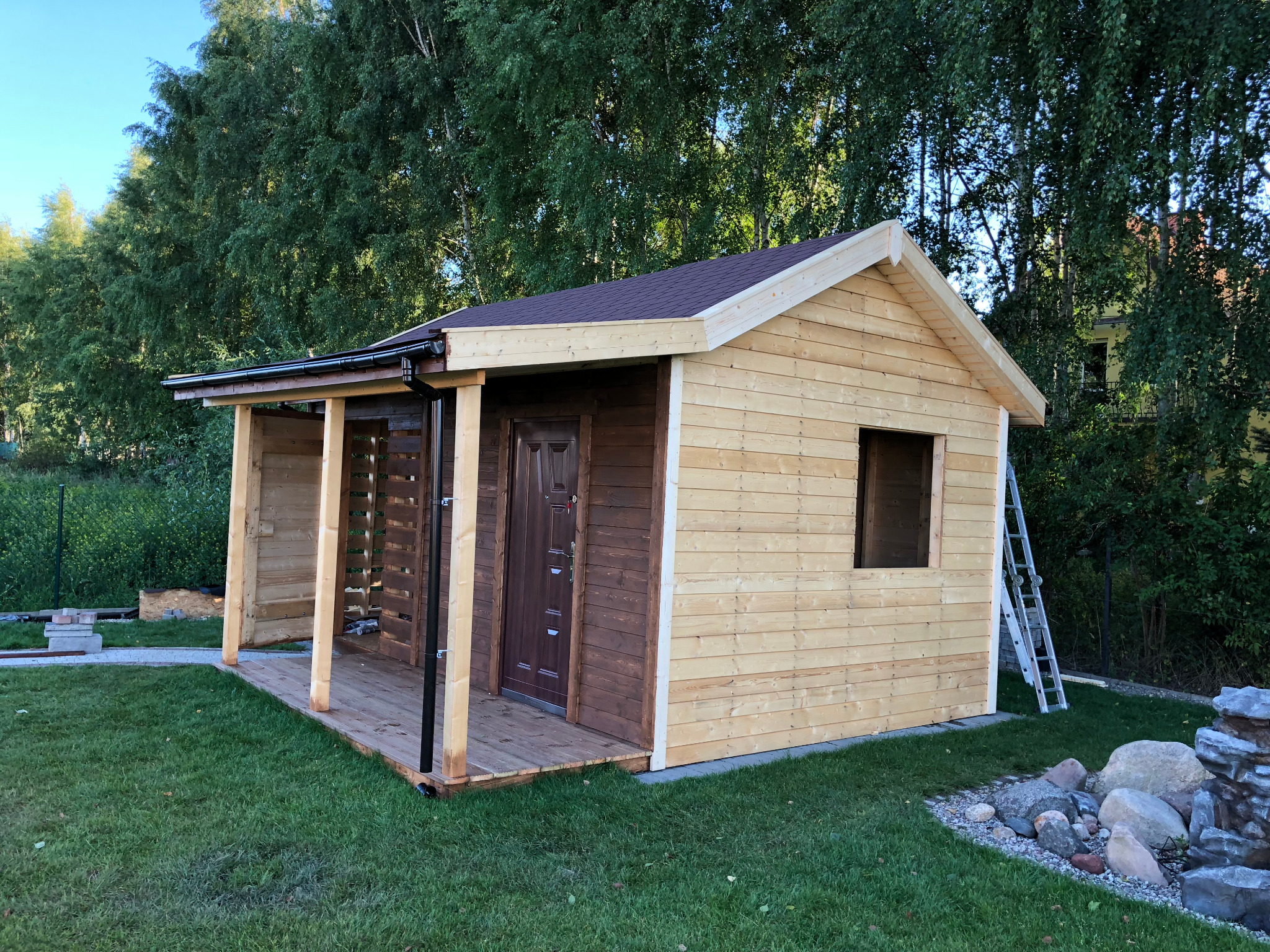 Drewniana altana ogrodowa z zadaszonym tarasem, częściowo pomalowana, z drabiną opartą o ścianę, na tle zielonego trawnika i drzew