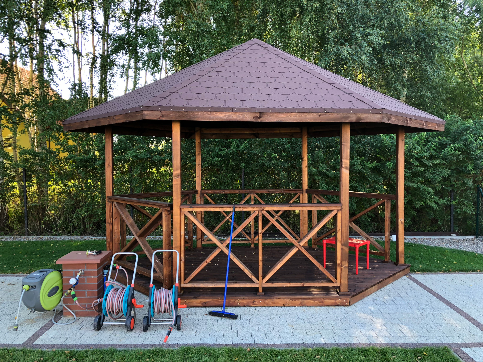 Ośmiokątna drewniana altana z brązowym dachem bitumicznym, widok z zewnątrz, obok zwinięte węże ogrodowe i czerwony stołek wewnątrz, niebieski mop oparty o balustradę