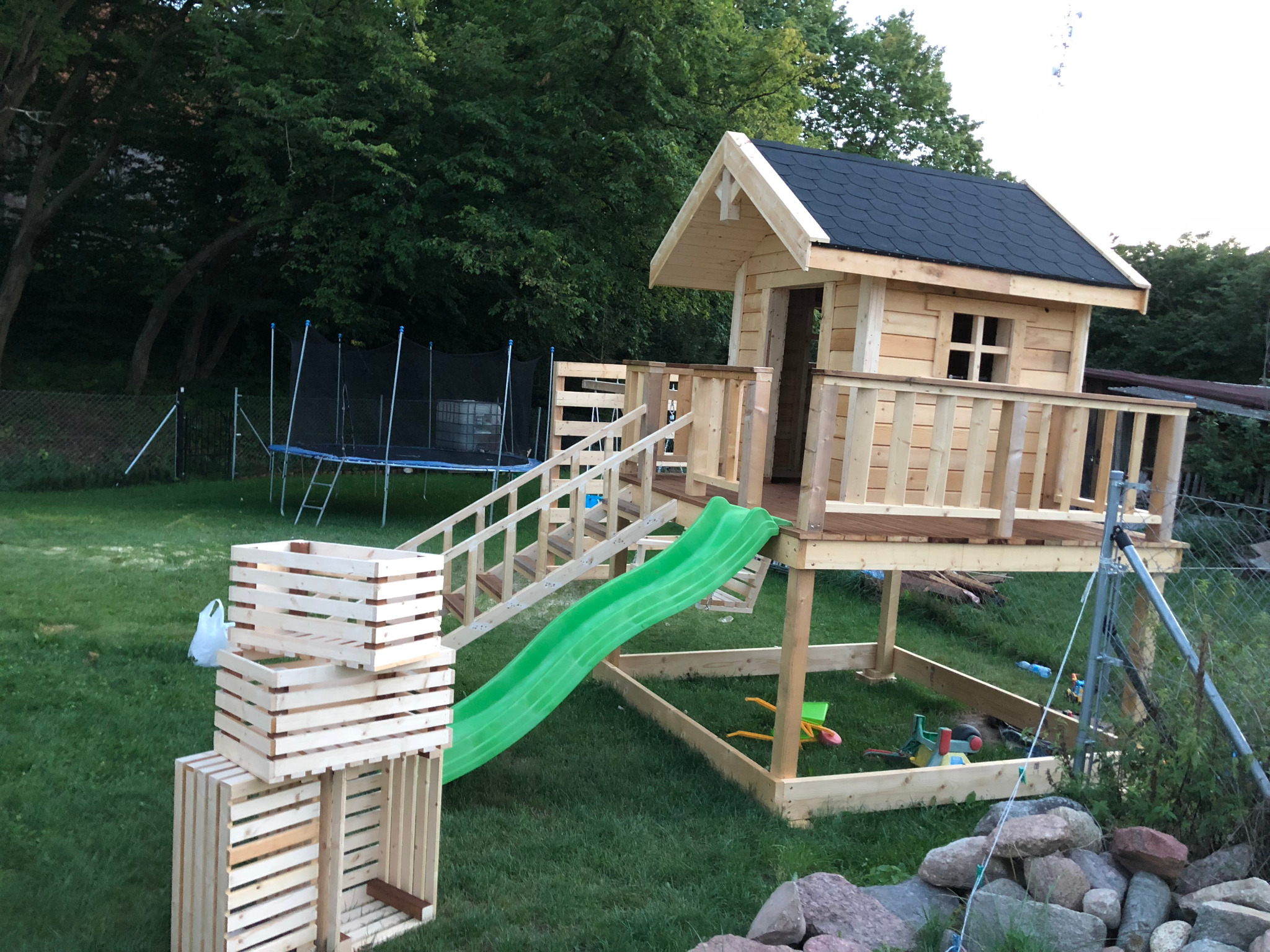Drewniany plac zabaw z domkiem na palach, zjeżdżalnią i konstrukcją z drewnianych skrzynek, umieszczony na trawniku z widocznym trampoliną w tle.