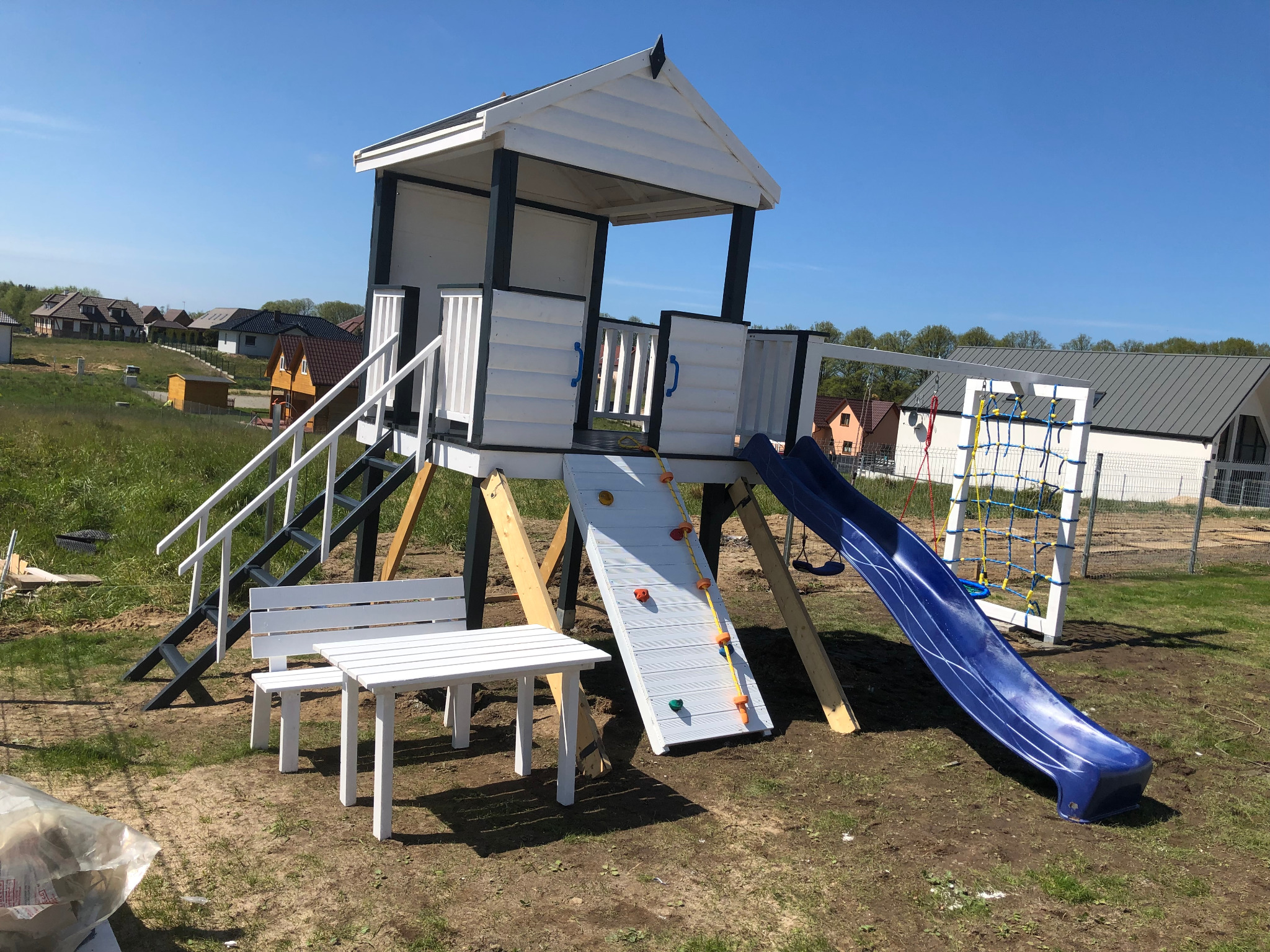 Drewniany plac zabaw dla dzieci z domkiem, zjeżdżalnią, ścianką wspinaczkową i stolikiem z ławeczką, na tle domów jednorodzinnych.