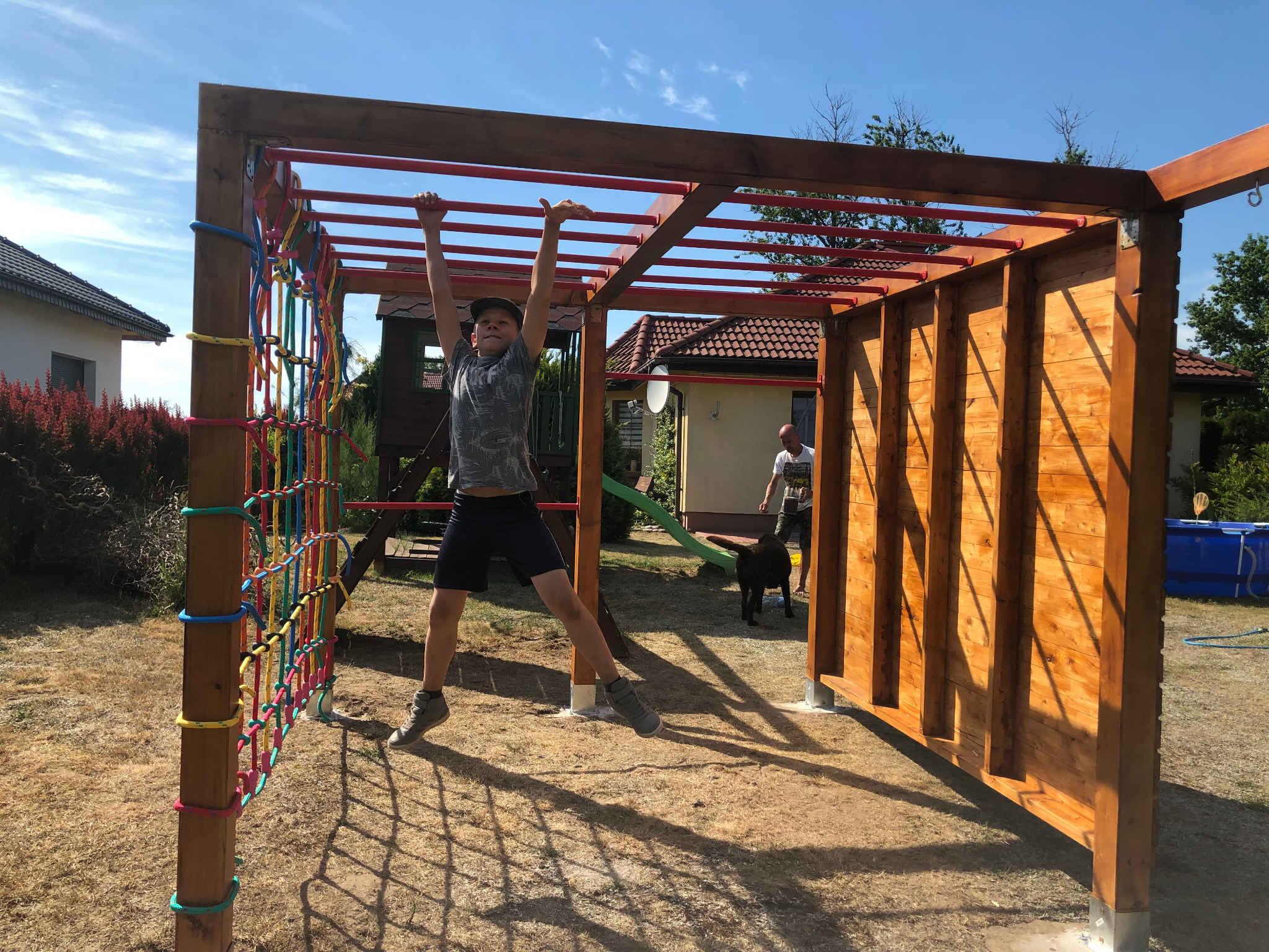 Chłopiec wisi na drewnianej konstrukcji z drabinkami i siatką w ogrodzie z domami w tle, w słoneczny dzień.