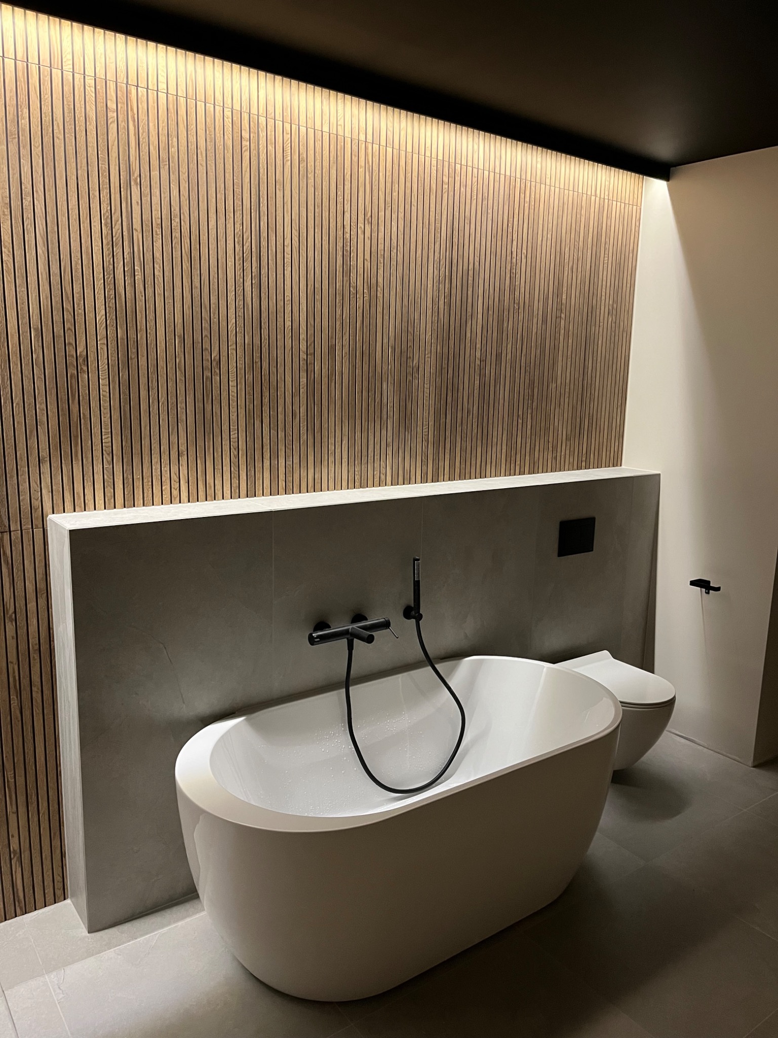 Nowoczesna łazienka z wolnostojącą białą wanną, czarną armaturą i drewnianą ścianą z podświetleniem LED, minimalistyczny design.