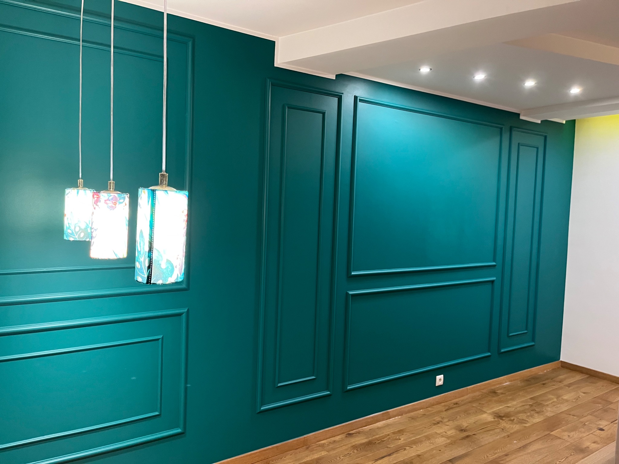 Turkusowa ściana z dekoracyjnymi panelami, drewniana podłoga i trzy wiszące lampy z motywem kwiatowym.