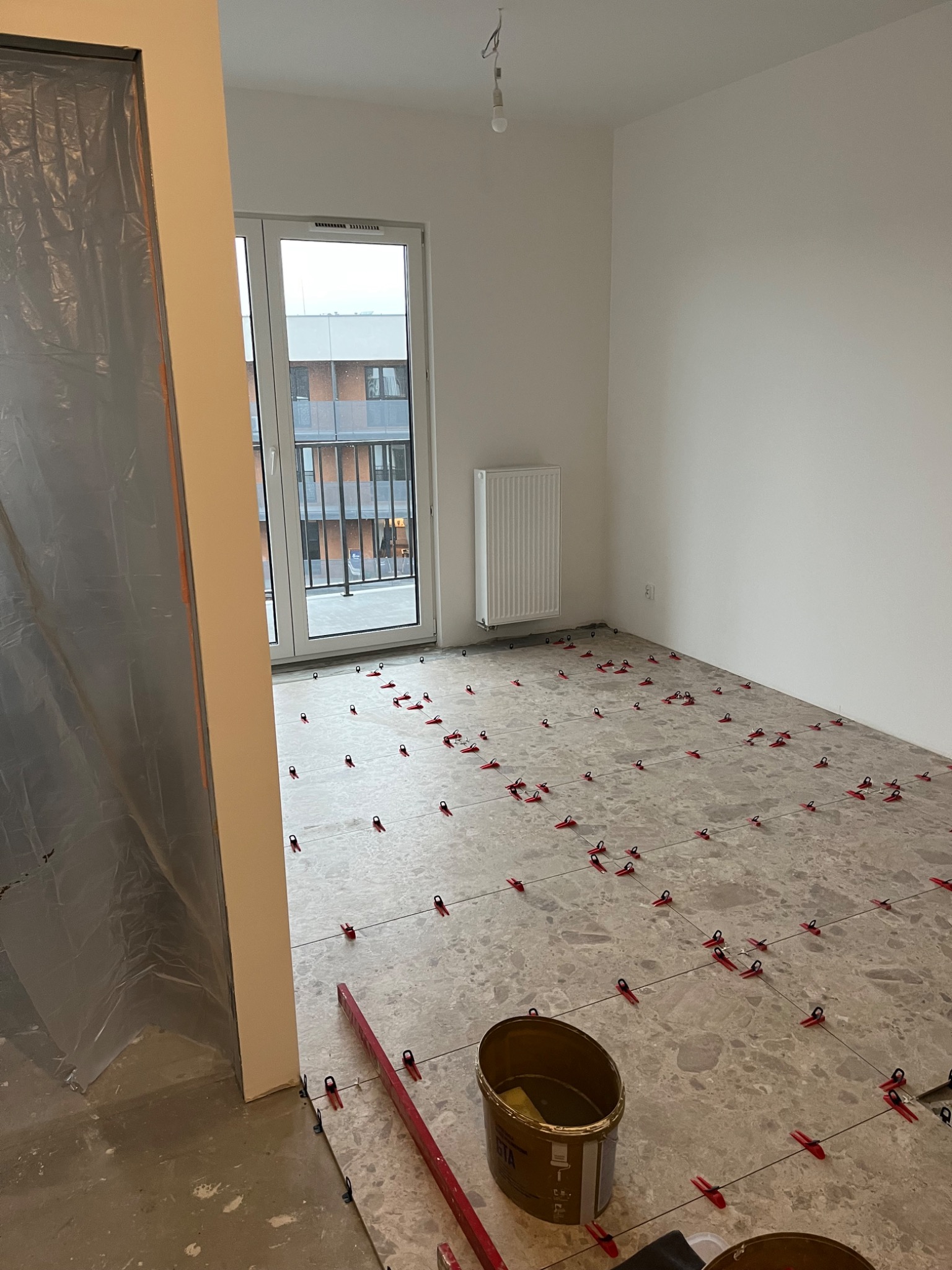 Układanie płytek podłogowych w pokoju, widoczne krzyżyki dystansowe, wiadro z klejem, poziomica i folia ochronna na ścianie