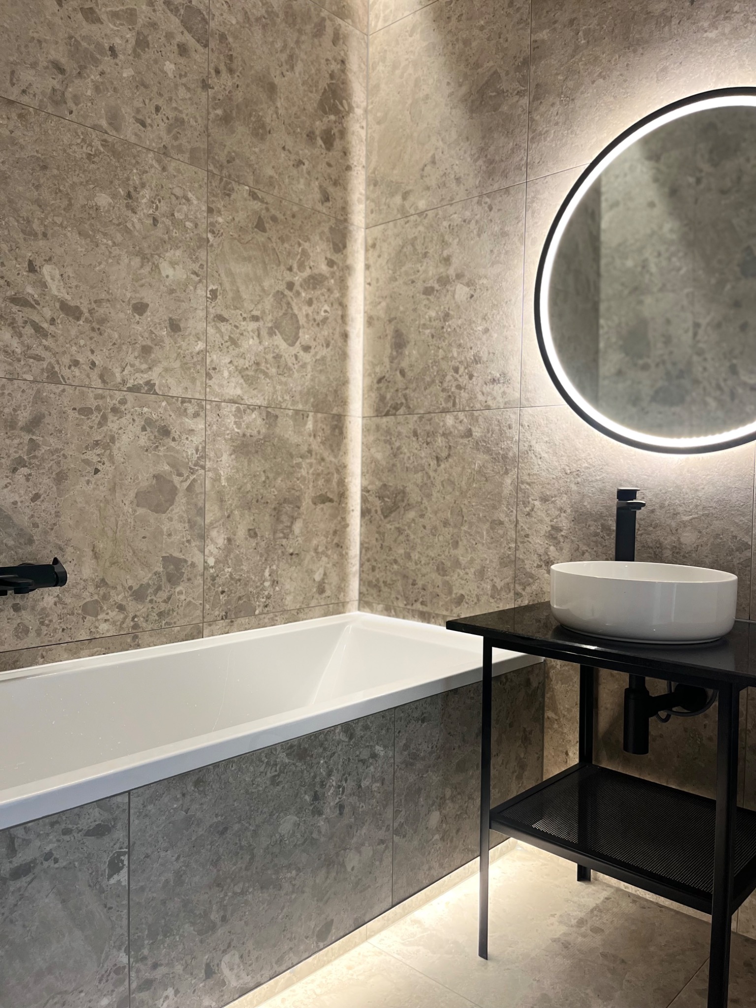 Nowoczesna łazienka z wanną obudowaną płytkami imitującymi kamień, okrągłym lustrem z podświetleniem LED i umywalką nablatową na czarnym metalowym stelażu.