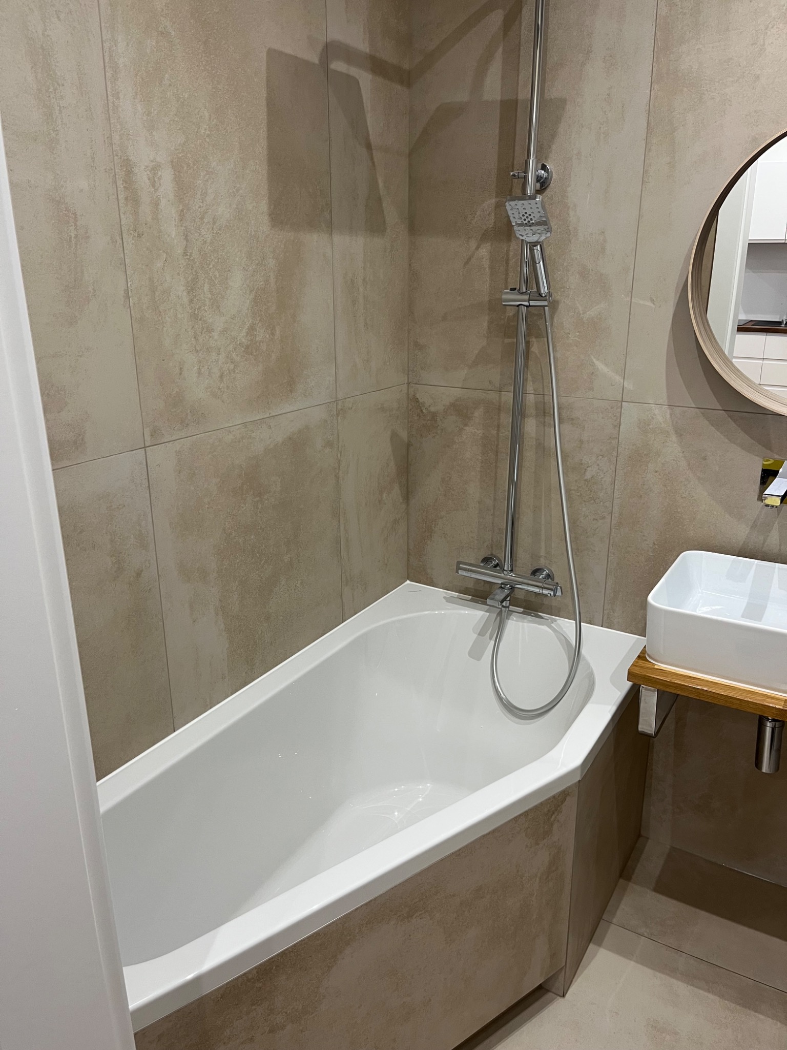 Nowoczesna łazienka z beżowymi płytkami imitującymi kamień, biała wanna z panelem, minimalistyczna umywalka na drewnianym blacie i okrągłe lustro.