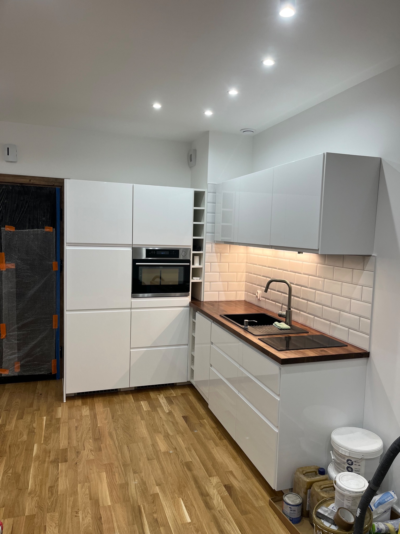 Nowoczesna kuchnia z białymi szafkami, drewnianym blatem i białymi płytkami typu metro. Na podłodze parkiet. Widoczne pojemniki z farbą i odkurzacz.