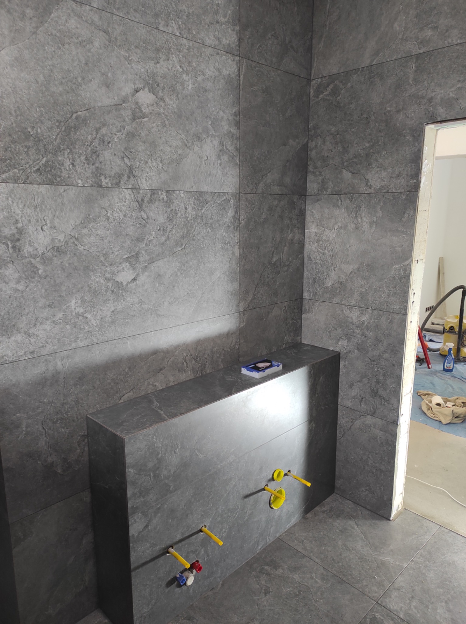 Szare płytki imitujące kamień na ścianach i podłodze, zabudowa stelaża WC z widocznymi przyłączami wodnymi, częściowo wykończona łazienka w trakcie prac.